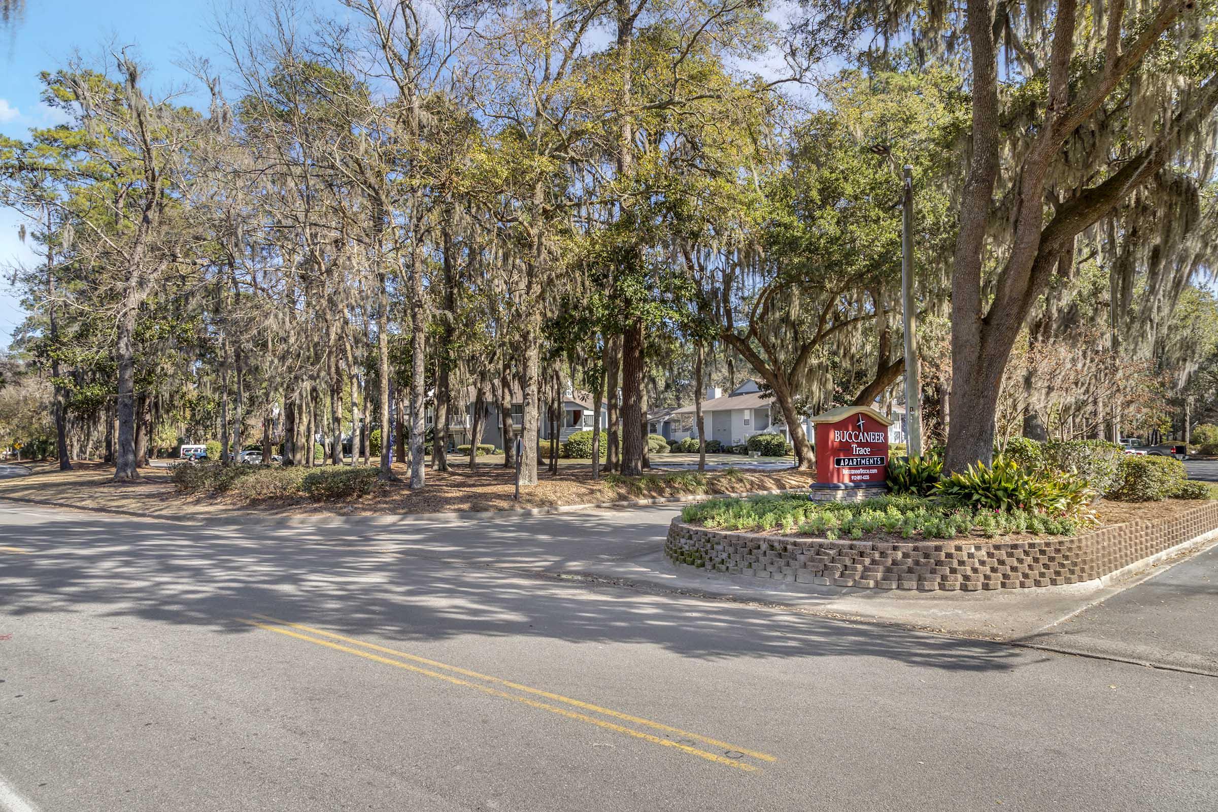 A view of a tree-lined street with a sign for a community entrance. The area features lush greenery and a mix of trees, typical of a suburban setting. The sun is shining, and there are residential buildings visible in the background, surrounded by nature.
