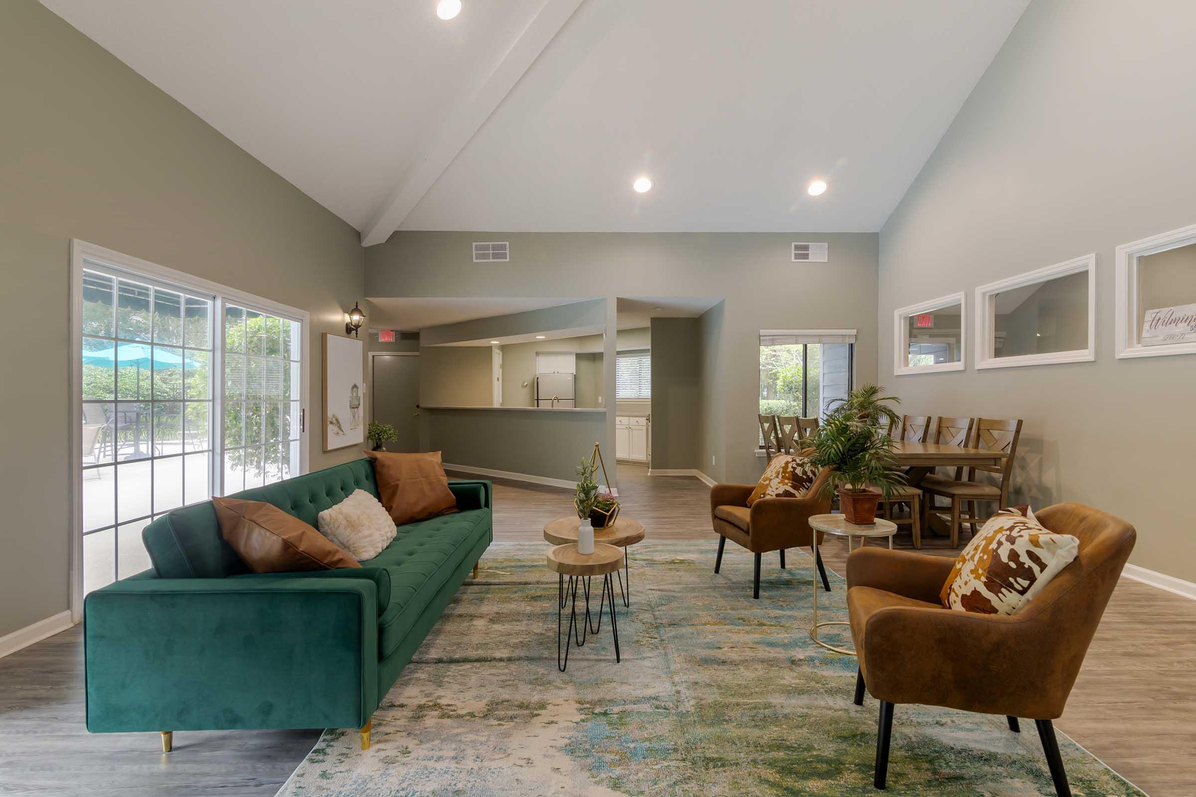 Interior of a modern living space featuring a green sofa with cushions, two brown chairs, a small round table, and a decorative plant. The room has vaulted ceilings, large windows letting in natural light, and neutral-colored walls. A dining area with a table and chairs is visible in the background.