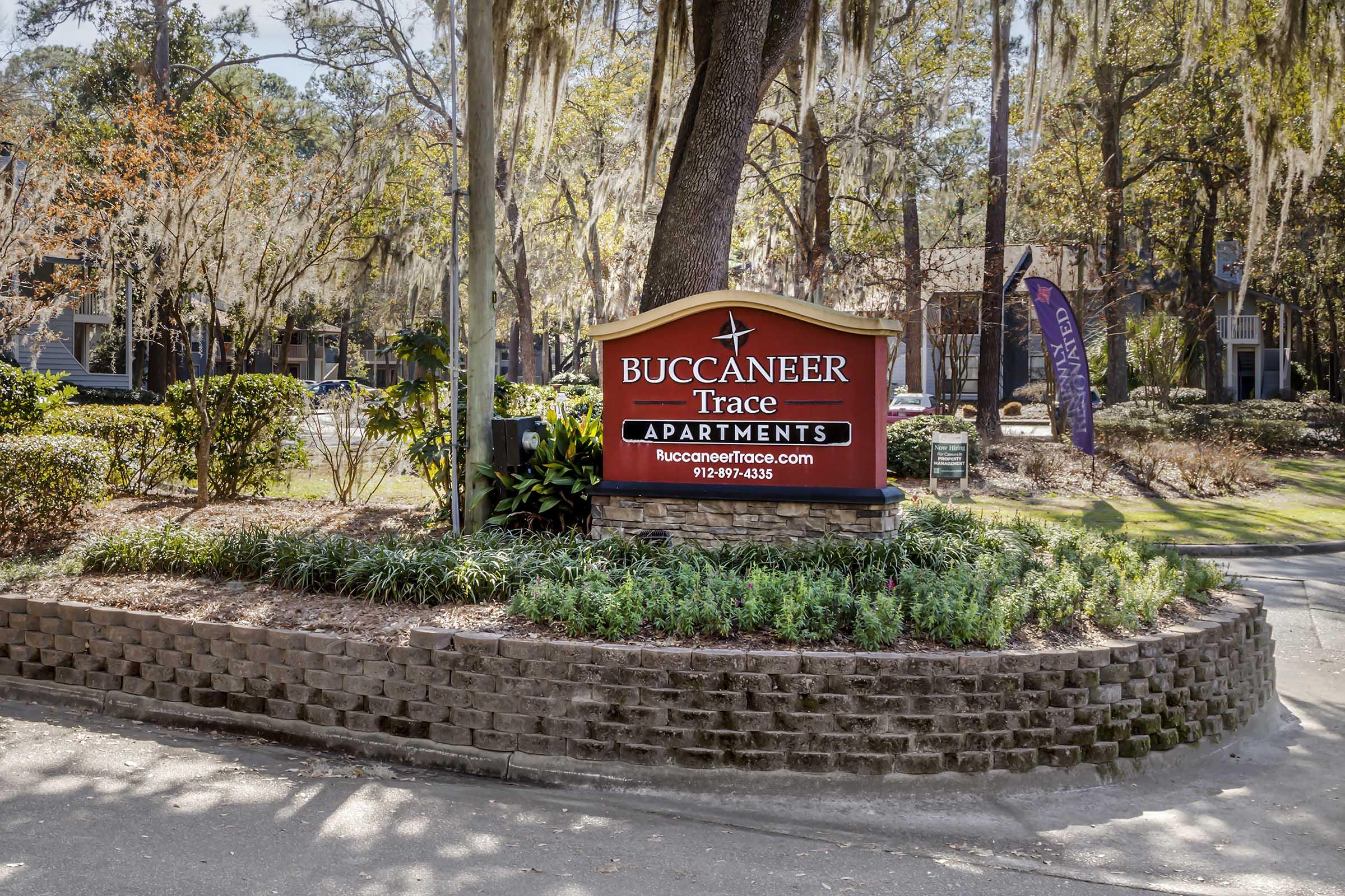Sign for Buccaneer Trace Apartments with a welcoming design, featuring a large logo and website information. Surrounded by landscaped greenery, the entrance presents a cozy, inviting atmosphere with trees and decorative plants. A purple banner is visible in the background.