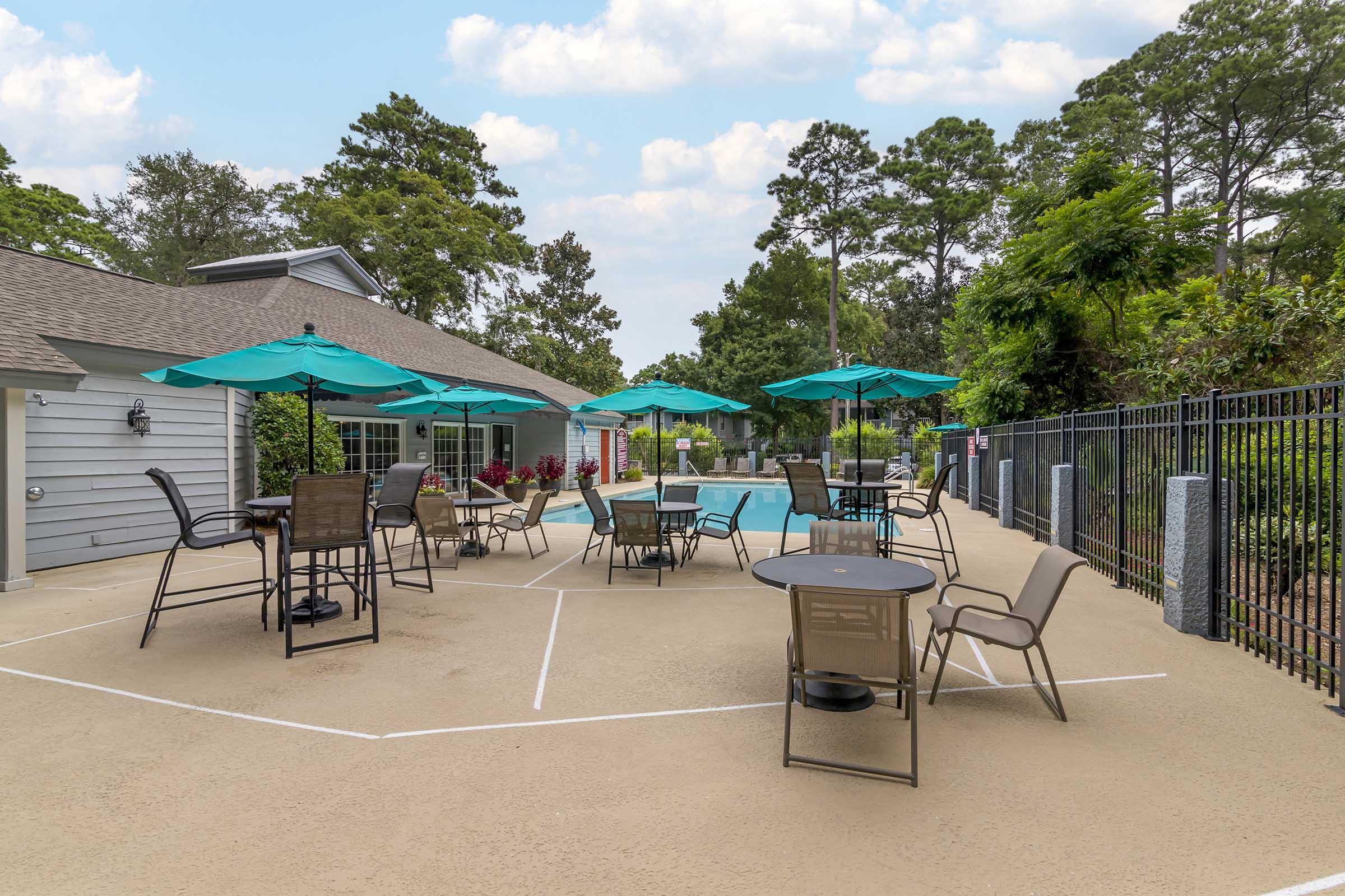 Outdoor pool area featuring lounge chairs and tables with teal umbrellas. Surrounded by greenery, the space includes a pool and a nearby clubhouse, with a fenced perimeter for privacy. Bright sky and trees create a relaxing atmosphere.