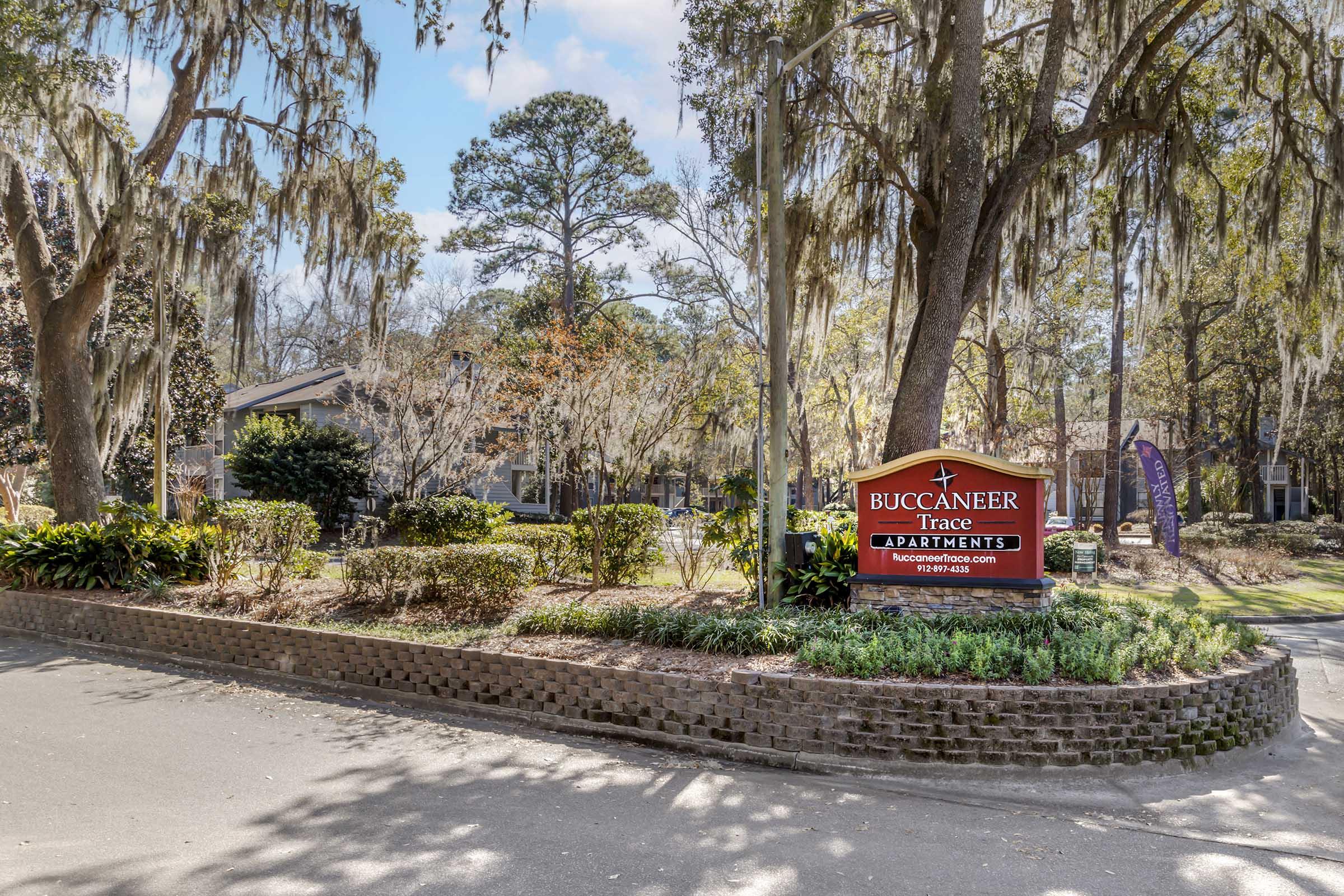 Sign for Buccaneer Trace Apartments, surrounded by lush greenery and trees with Spanish moss, set in a peaceful residential area. The sign is prominently displayed at the entrance, welcoming visitors and residents. Clear blue sky and natural landscaping enhance the inviting atmosphere of the property.