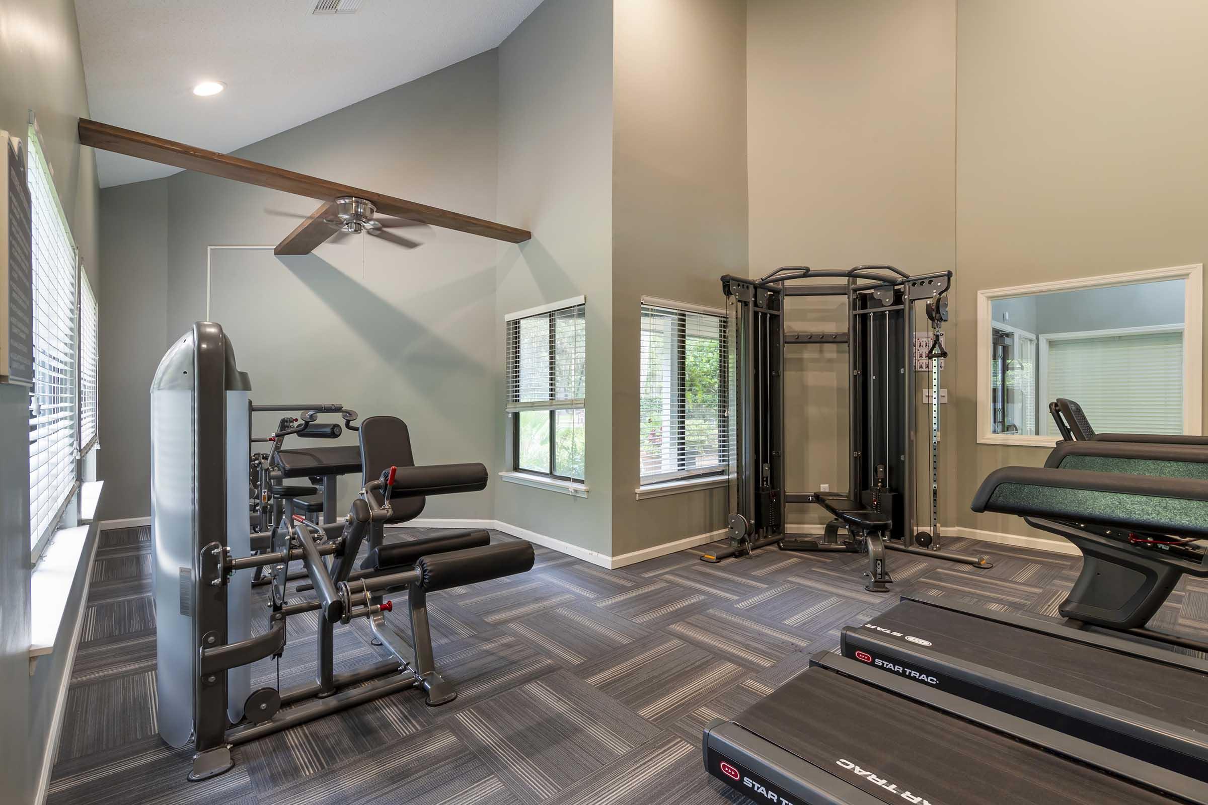 A modern fitness room featuring various gym equipment, including a weight machine, exercise bench, and cardio machines. Large windows provide natural light, and the room has a neutral color palette with textured flooring. A ceiling fan is visible in the upper corner.
