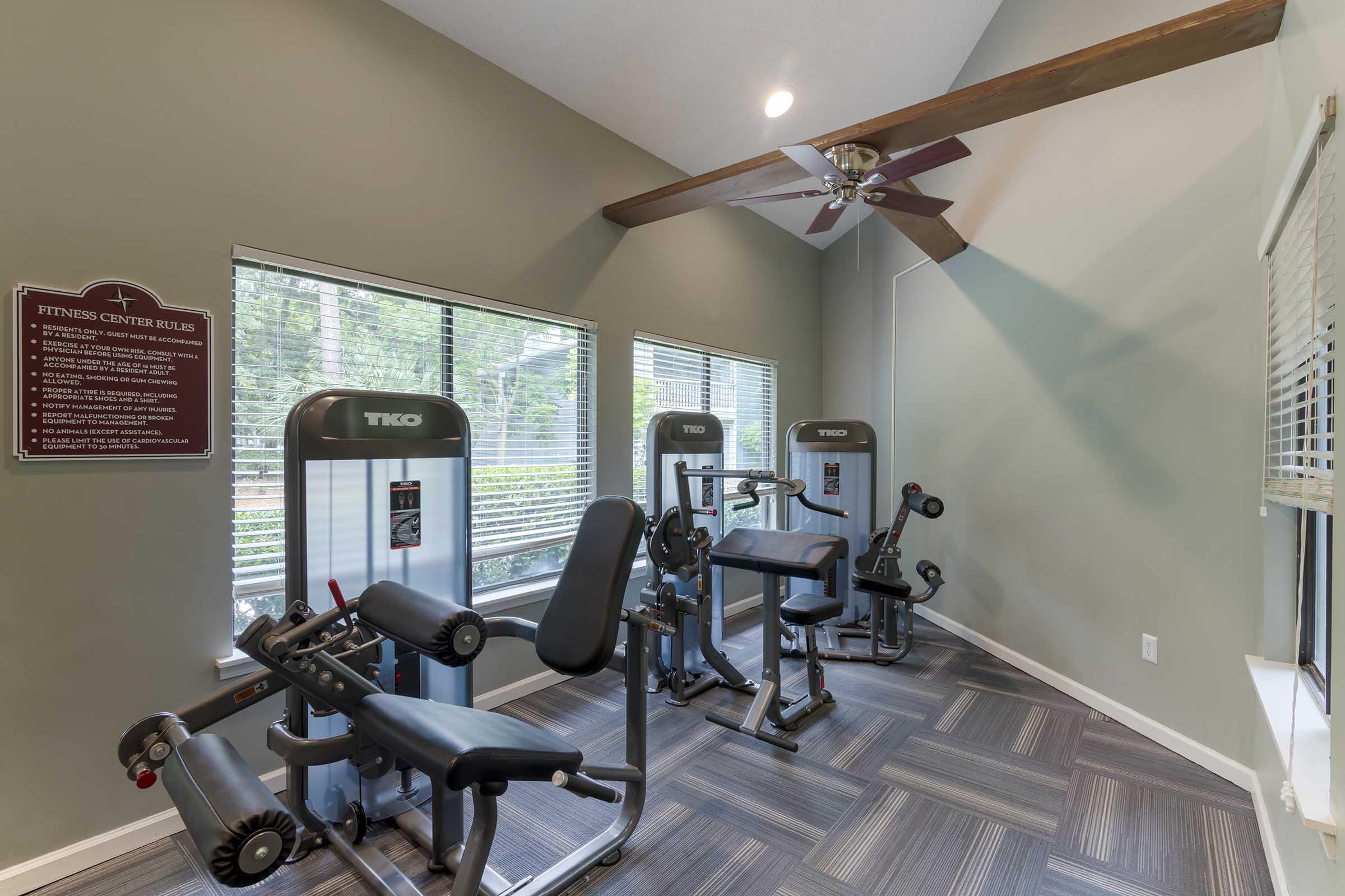 A fitness center with exercise equipment, including weight machines and benches. Large windows allow natural light to enter the room, and a ceiling fan is visible above. Fitness center rules are displayed on a sign on the wall, and the flooring features a patterned design.