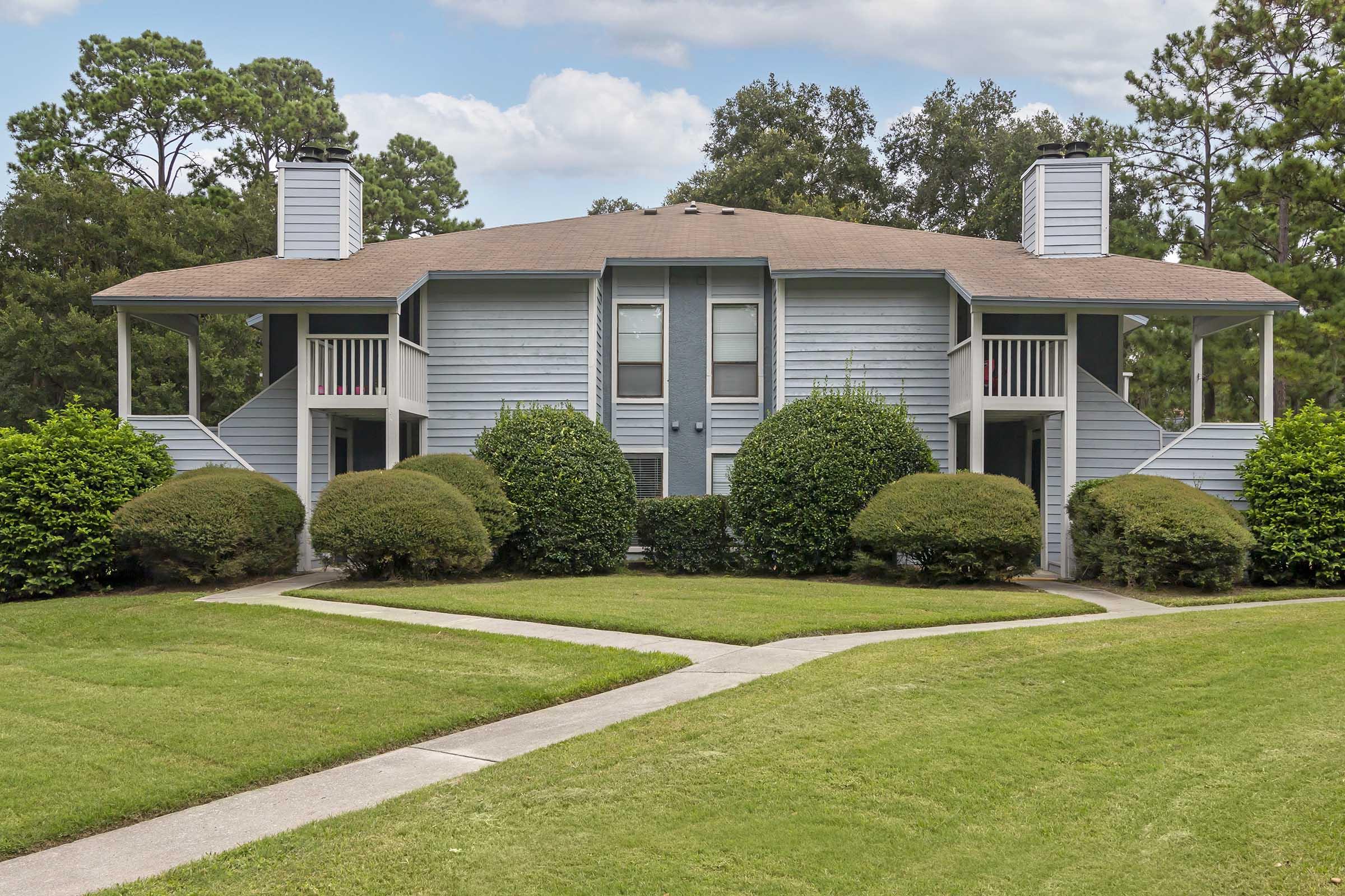 A two-story apartment building with a light blue exterior, featuring two entrances on the front and surrounded by neatly trimmed bushes and landscaping. A concrete pathway leads up to the building, which is set against a backdrop of trees and a cloudy sky.