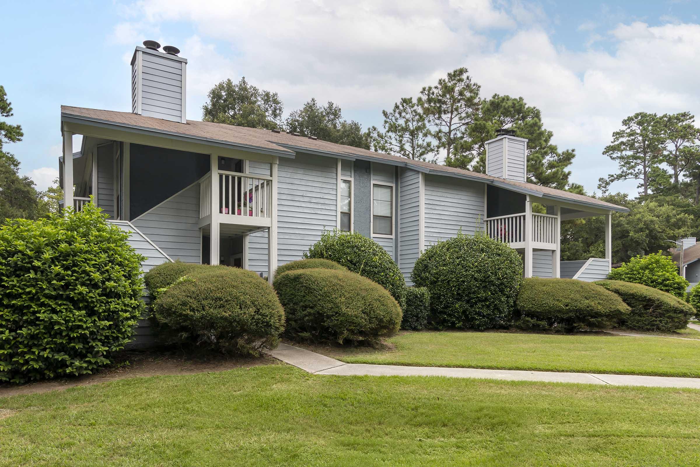 A two-story residential building with a light blue exterior and multiple balconies. The architecture features gray accents and two chimneys. Lush green shrubs line the pathway leading to the entrance, surrounded by a well-maintained lawn and trees in the background under a partly cloudy sky.