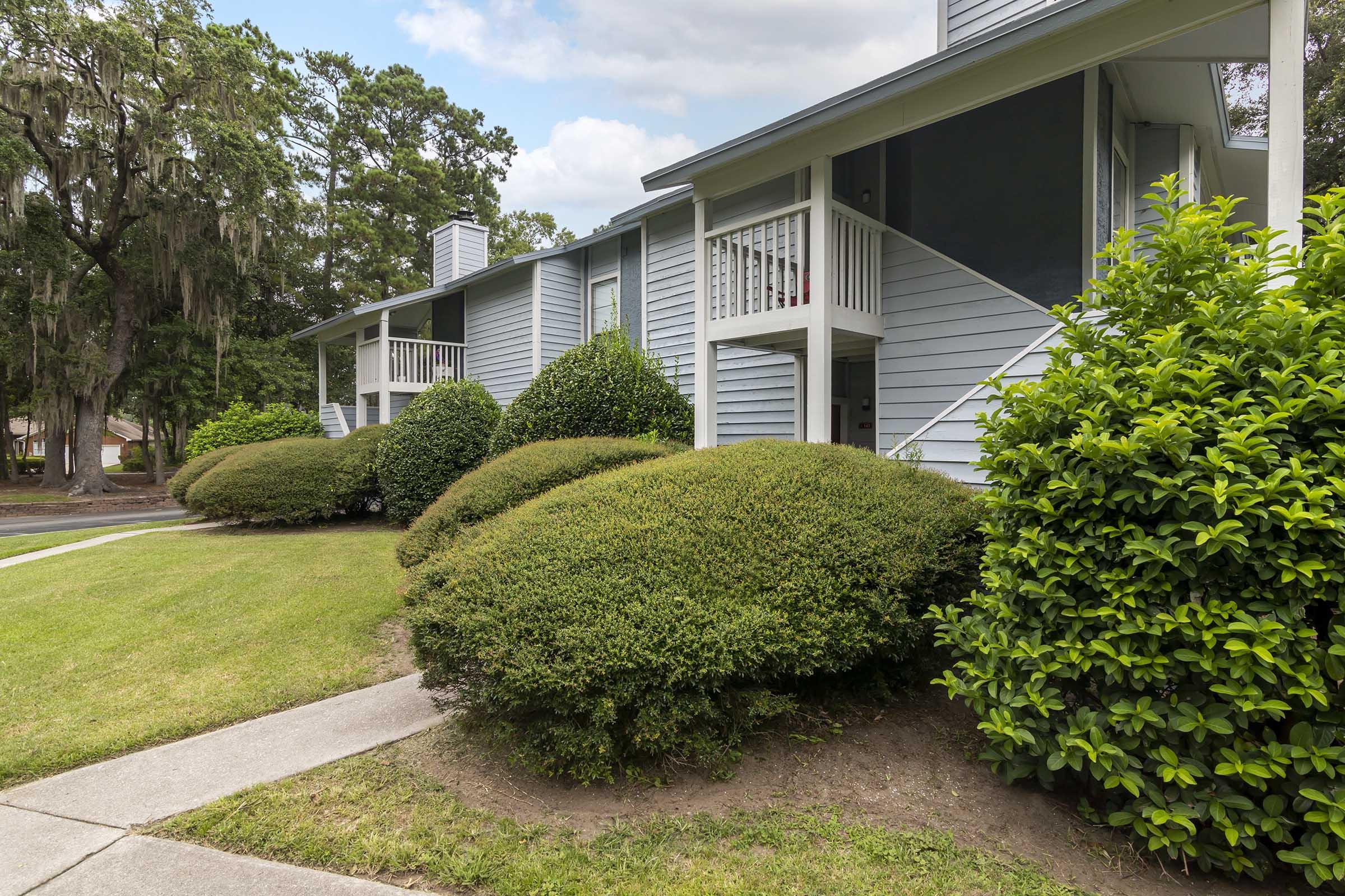 Two-story apartment building with a light blue exterior, featuring white balconies and railings. The building is surrounded by well-manicured shrubs and lawns, with a concrete pathway leading towards it. Trees in the background provide a natural setting, and the sky is partly cloudy.