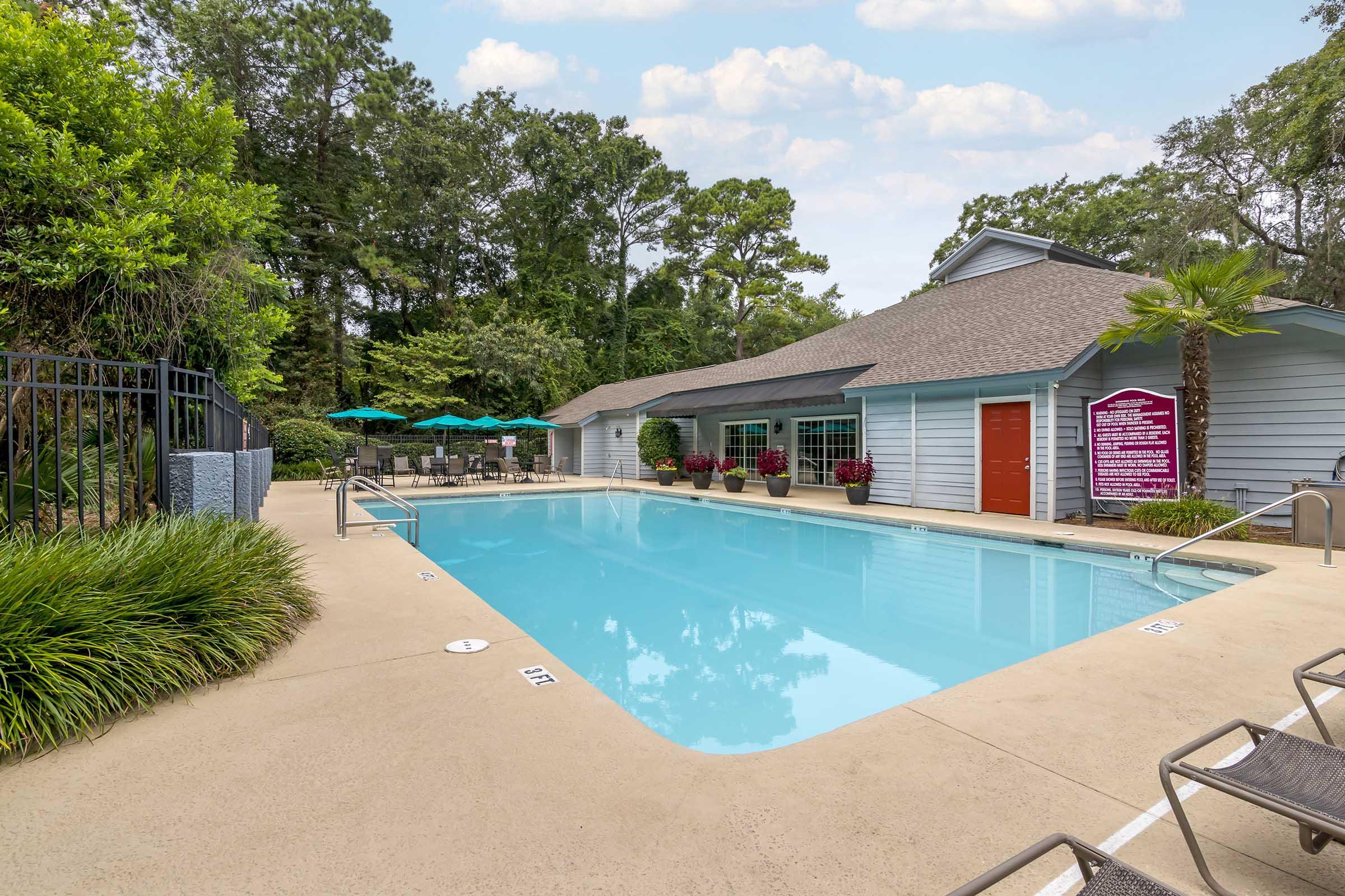 A clear swimming pool surrounded by green trees and shrubs. Lounge chairs are positioned nearby, and there are umbrellas set up for shade. A building with a red door and a sign is visible in the background, enhancing the inviting atmosphere of the space.
