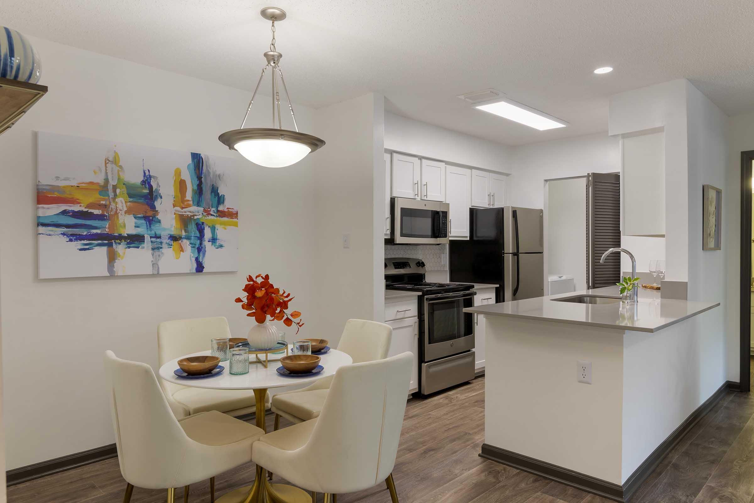 A modern kitchen and dining area featuring a round table with white chairs, set with bowls and glasses. The kitchen includes stainless steel appliances and an island with a sink. A colorful abstract painting hangs on the wall, and a vase with red flowers adds a pop of color to the dining table.