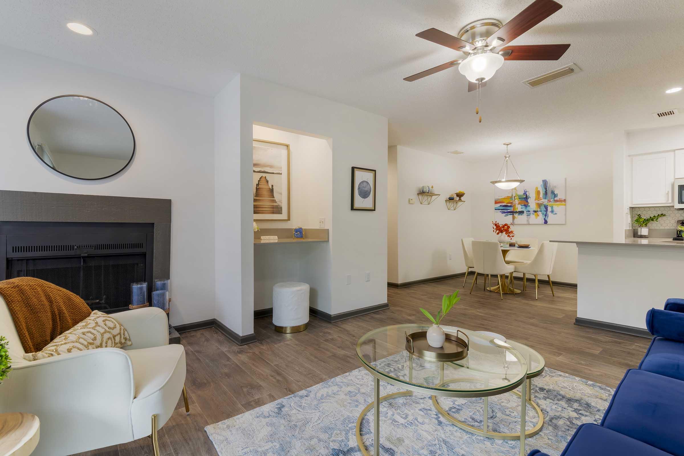 A cozy living room featuring a white armchair with a brown throw, a glass coffee table on a blue area rug, and a blue sofa. There's a fireplace, a round wall mirror, and a small dining area visible in the background with a pendant light and decorative artwork.