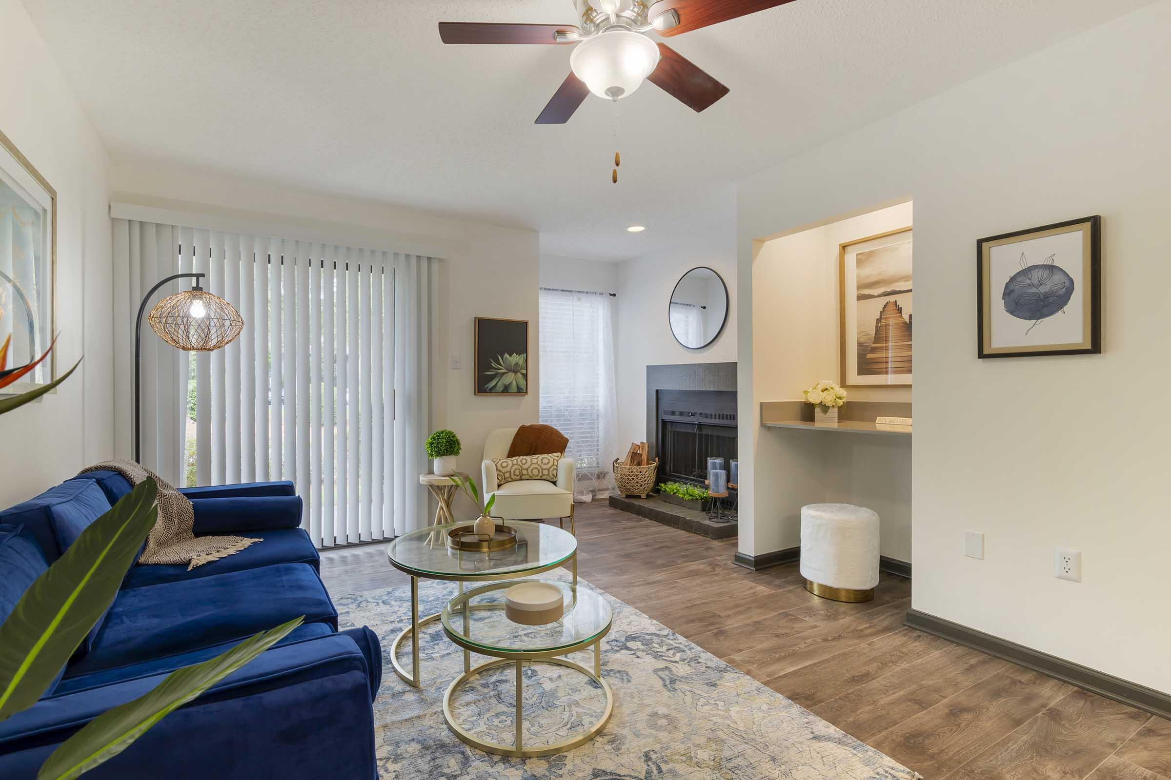 A modern living room featuring a blue sofa, round glass coffee table, and stylish decor. The room has a ceiling fan, large windows with vertical blinds, and a cozy reading chair. A fireplace is visible in the background, with art and plants enhancing the inviting atmosphere.
