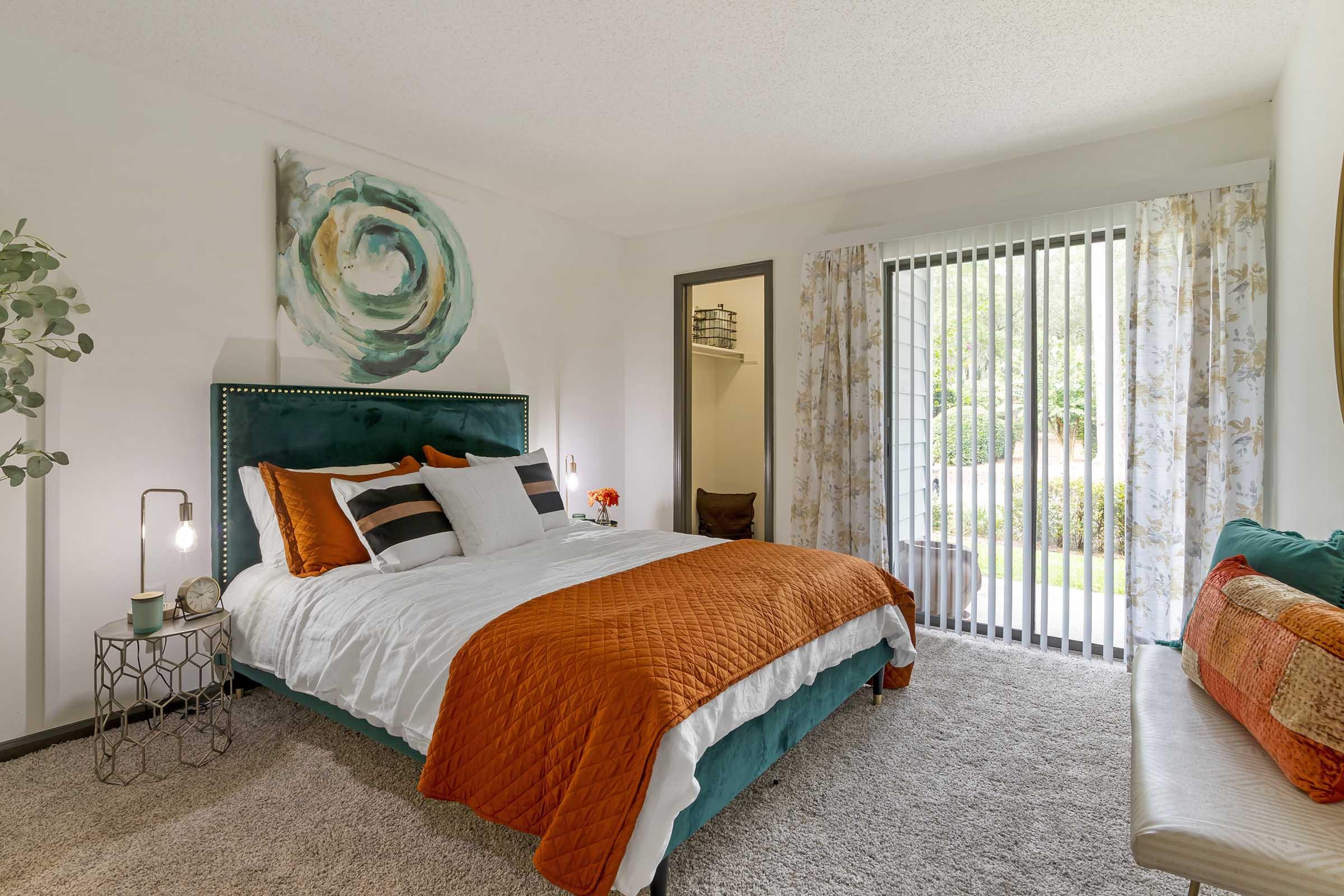 A cozy bedroom featuring a green and white color scheme with an orange quilt and decorative pillows. A large window with vertical blinds allows natural light, while a stylish lamp and a small table add elegance. A piece of abstract wall art complements the décor, and a plush seating area enhances comfort.