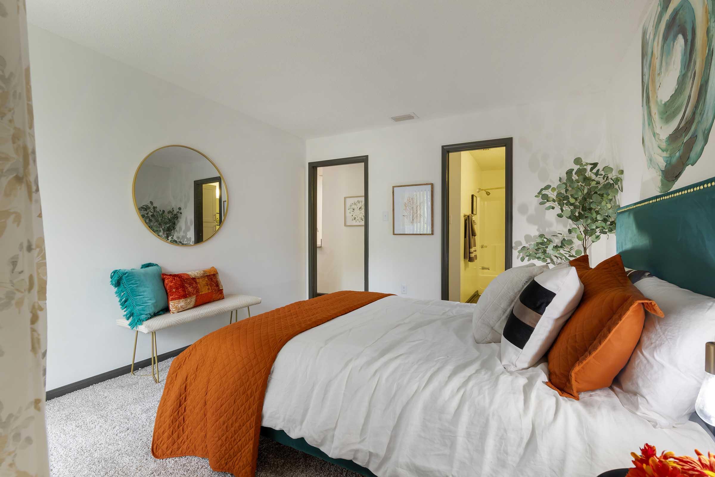 Cozy bedroom featuring a plush bed with orange and white bedding, decorative pillows, and a stylish round mirror on the wall. The room includes a bench with teal and white pillows, and a soft carpet, with a door leading to a bathroom visible in the background. Bright and inviting atmosphere.