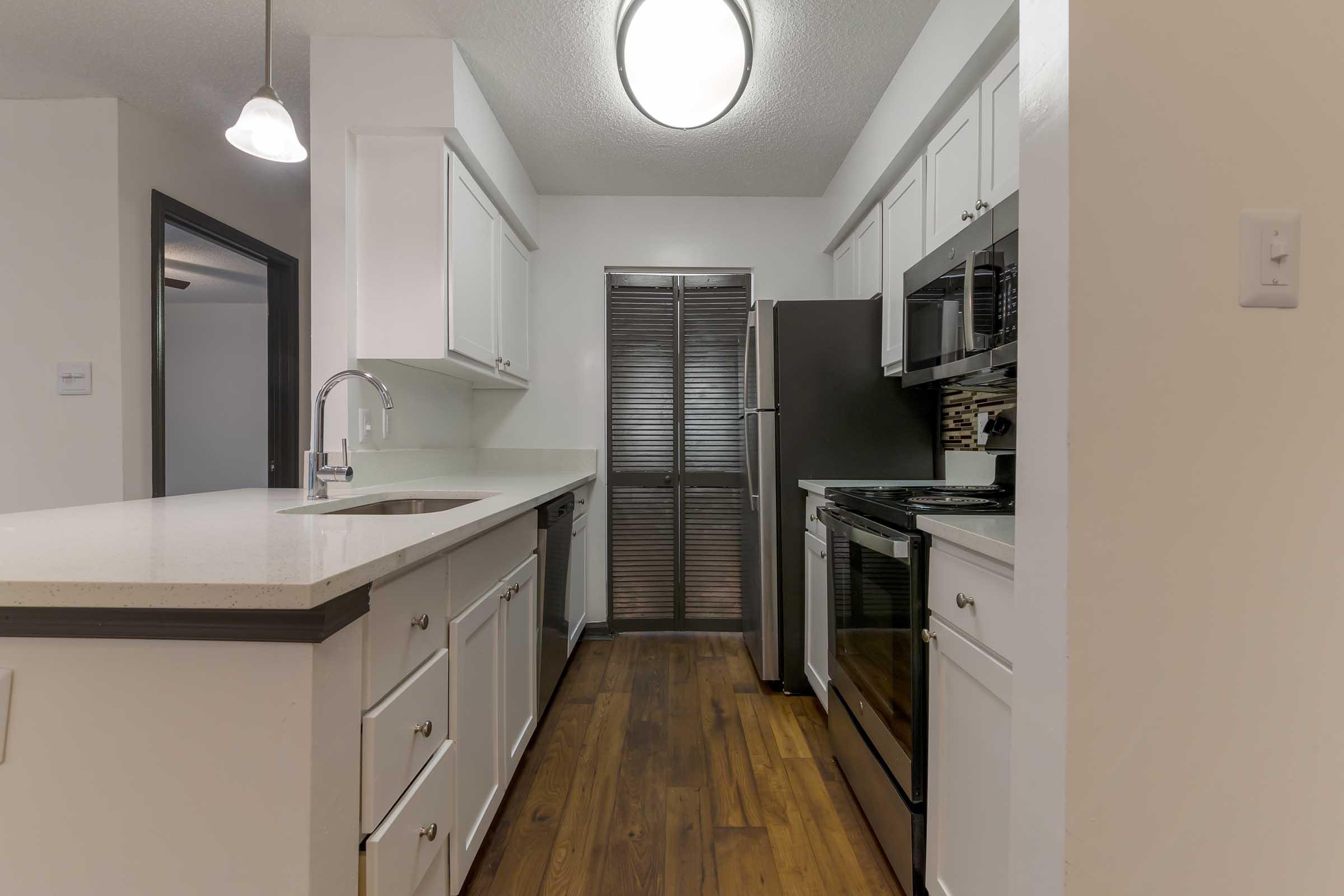 Modern kitchen featuring white cabinets, a quartz countertop, stainless steel appliances including an oven, microwave, and refrigerator, as well as a sleek sink. The floor is made of warm wooden planks, and the space is well-lit with overhead lights. The design emphasizes a contemporary aesthetic with a functional layout.