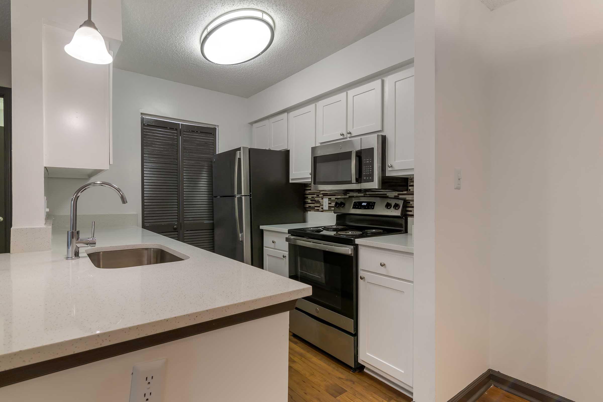 Modern kitchen featuring white cabinets, a stainless steel refrigerator, and a stainless steel stove with an oven. The countertops are light, and there's a sleek sink with a faucet. A light fixture hangs from the ceiling, and there is a window with blinds on the left, enhancing the bright atmosphere.