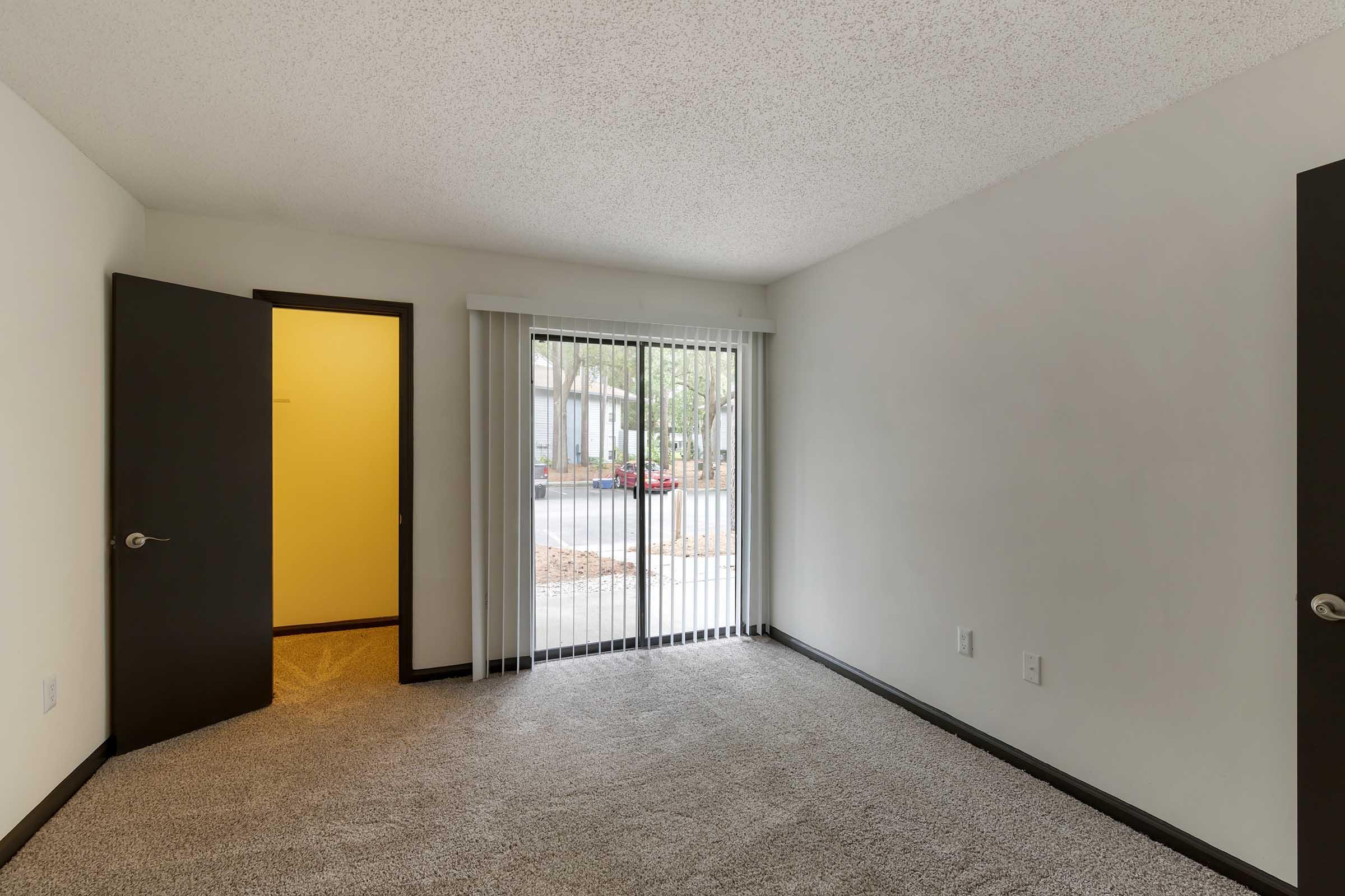 A well-lit empty room featuring light carpet, beige walls, and dark door frames. Two doors are visible: one leading outside with vertical blinds, and another open to a hallway. The space is clean and ready for furniture, giving a sense of potential for design options.