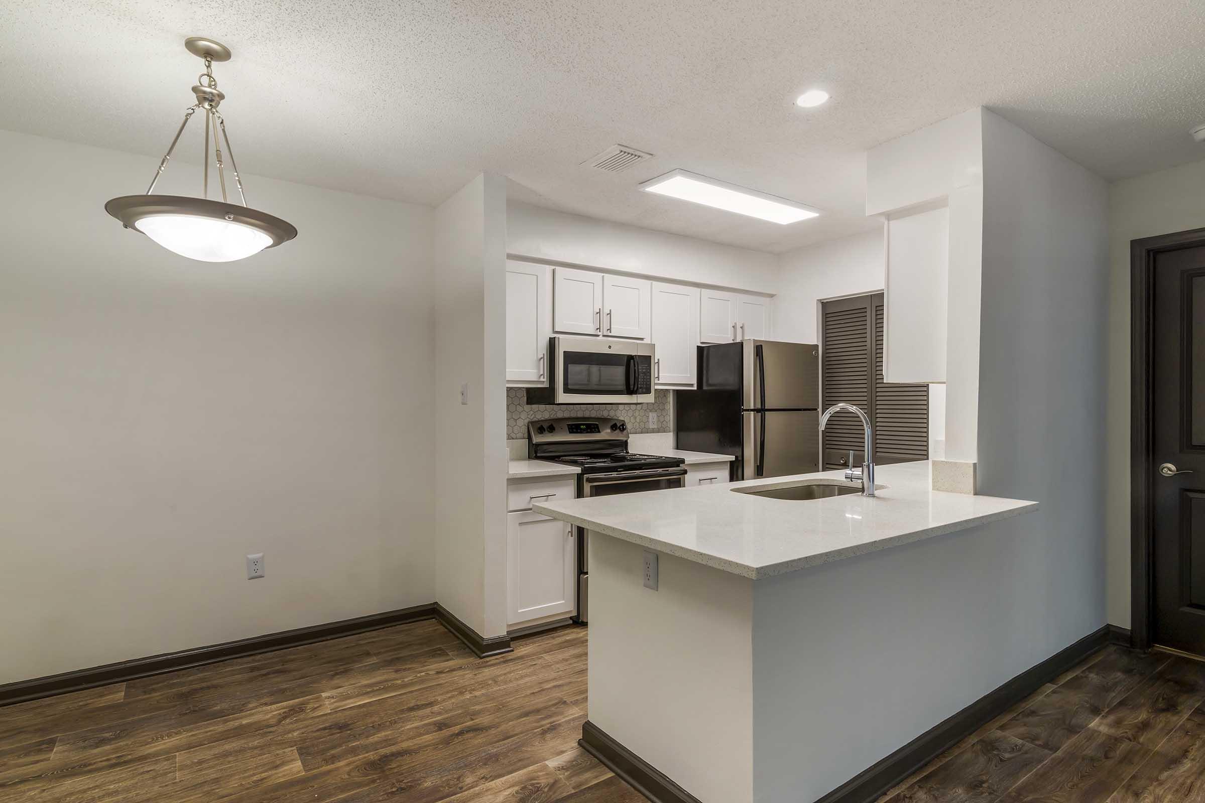Modern kitchen with white cabinetry, stainless steel appliances, and a spacious island. Features include a microwave, oven, and refrigerator. The flooring is a warm wood tone, and the lighting is bright, creating an inviting atmosphere. A pendant light hangs above the island, enhancing the space's appeal.