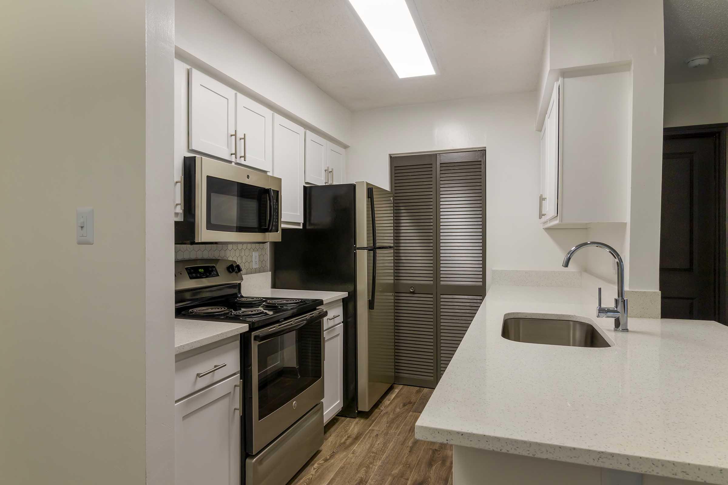 A modern kitchen featuring white cabinetry, stainless steel appliances including a double oven, microwave, and refrigerator. The countertop is a light-colored stone, and the kitchen includes a sink with a sleek faucet. A sliding door closet is visible in the background, enhancing storage options.