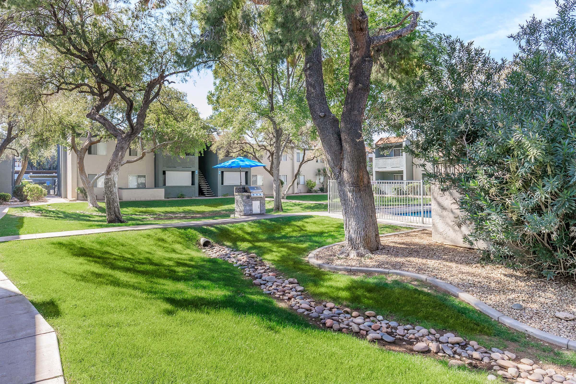 A landscaped area featuring lush green grass, walking paths, and trees. In the background, there are residential buildings with balconies. A blue umbrella shades a seating area near a pool protected by a fence. Decorative stones line a small pathway, enhancing the outdoor aesthetic.