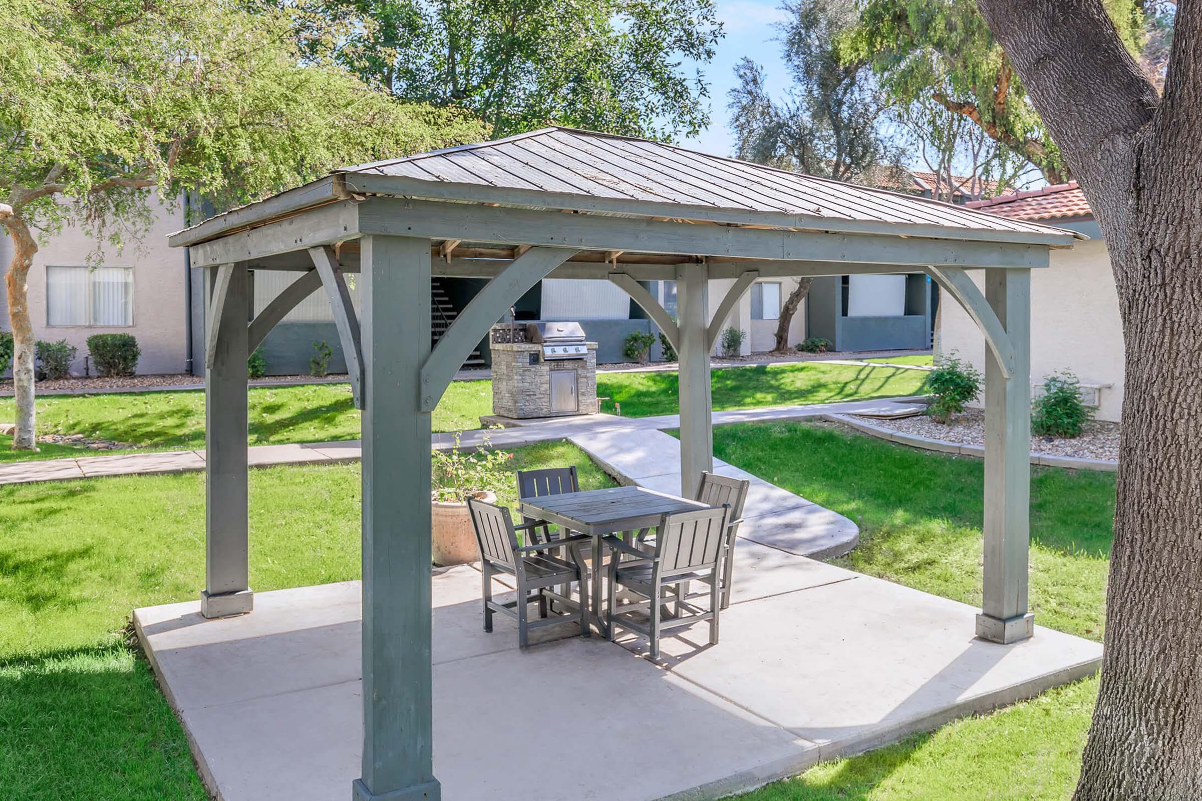 A wooden gazebo with a sloped roof sits on a concrete slab in a green lawn. Underneath, there is a table with four chairs. In the background, there are well-maintained apartment buildings and a barbecue grill, surrounded by trees and shrubs.