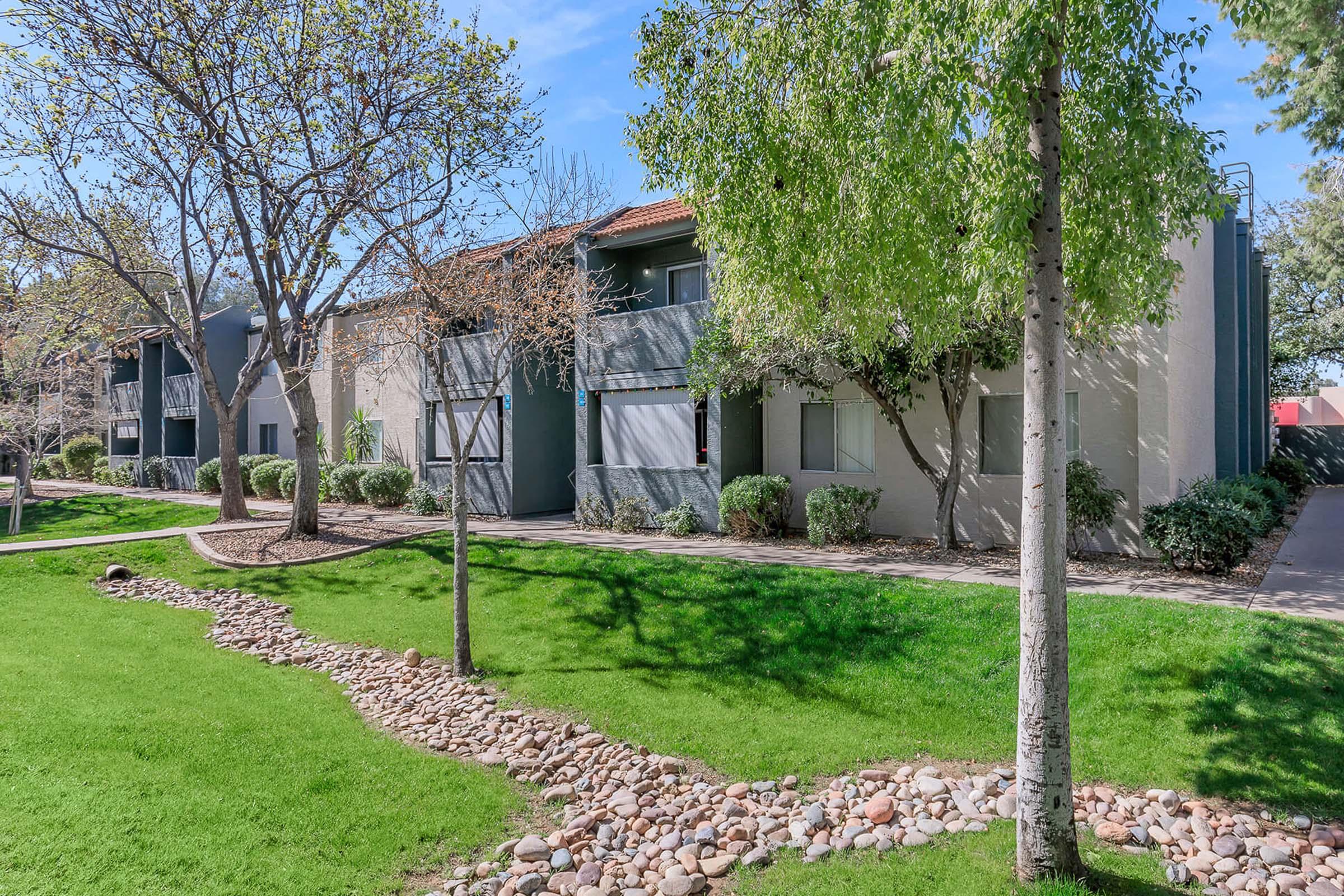 A vibrant landscape featuring a well-maintained apartment complex with green lawns and trees. The image shows two buildings with light-colored exteriors, surrounded by lush vegetation and a decorative rock path. Clear blue skies and sunlight enhance the inviting atmosphere of the area.