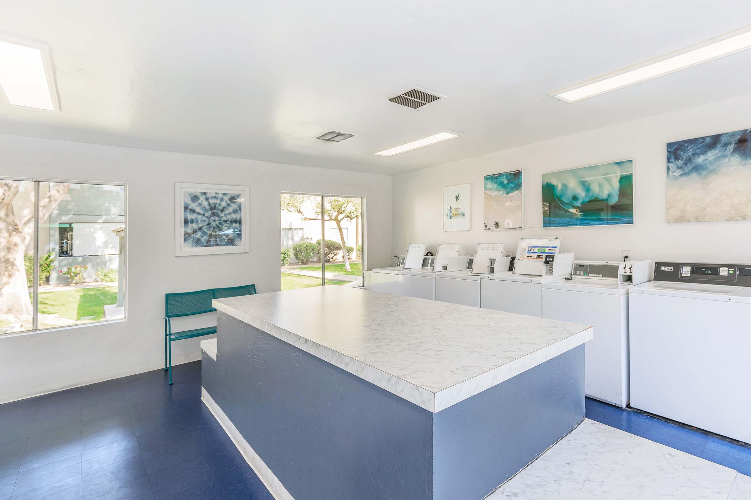A modern laundry room featuring white washing machines and dryers against a light-colored wall. There’s a large marble countertop in the center, and artwork hangs on the walls. The space is bright and well-lit, with windows overlooking a grassy area outside. A teal bench is positioned for seating.