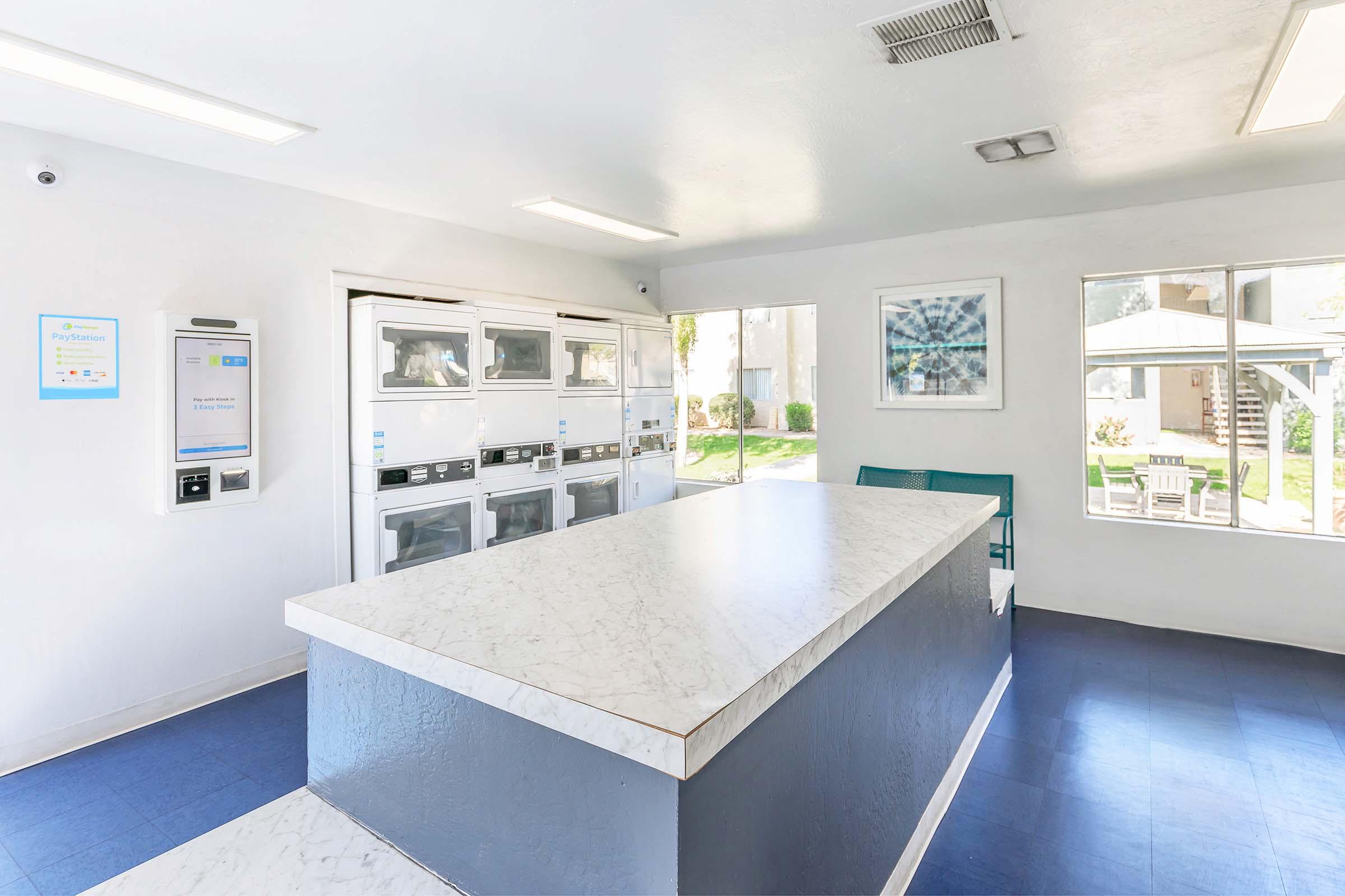 Bright laundry room featuring a large white table, several stacked washing machines and dryers, and a window with a view of the outdoors. The walls are painted white, and there are a couple of chairs for seating. A modern design is complemented by good lighting and a blue floor.