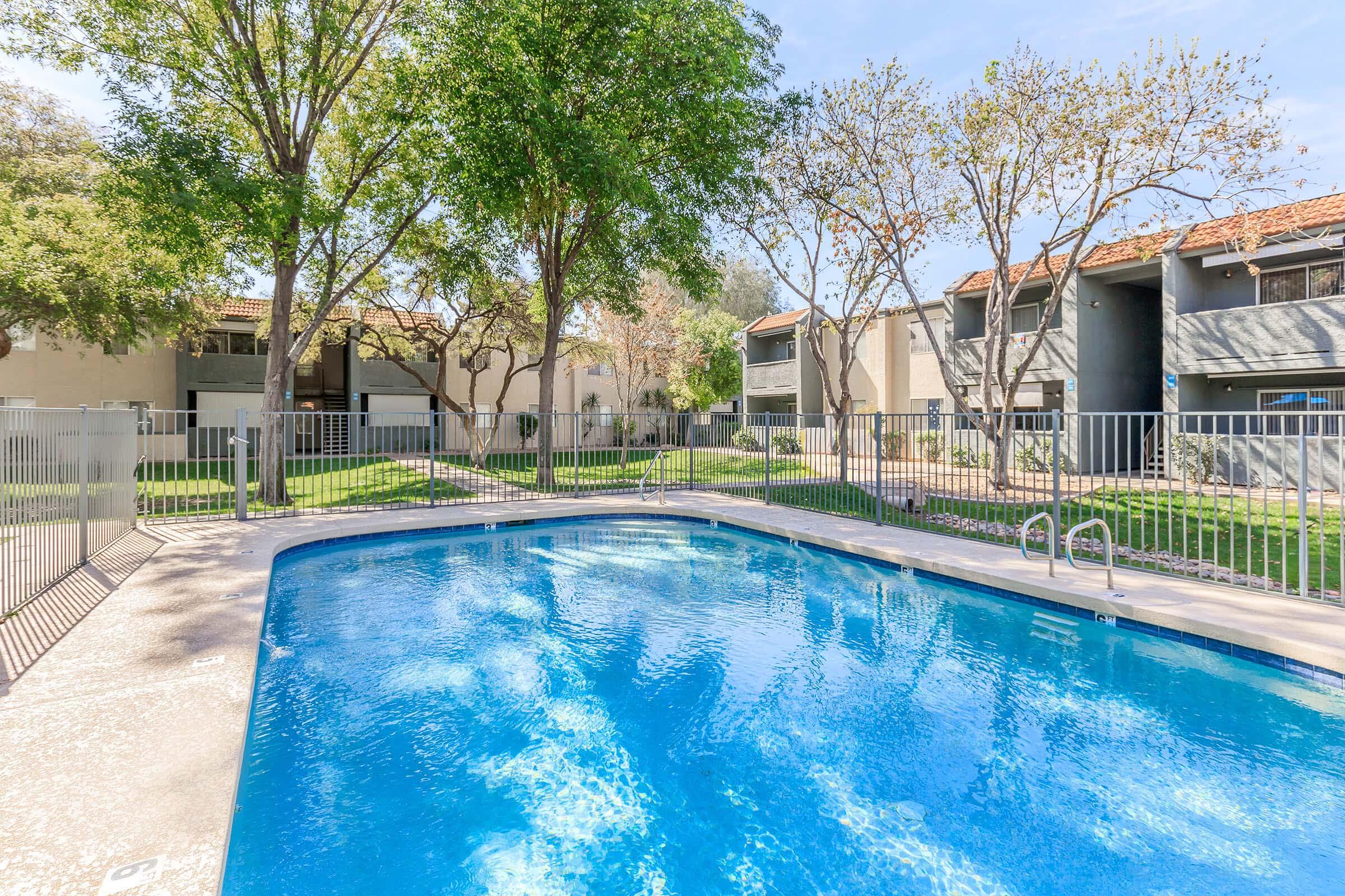 A clear swimming pool surrounded by a fence, with tall trees and green grass nearby. In the background, there are several apartment buildings with balconies, providing a serene outdoor environment. The sky is blue with a few clouds, indicating a pleasant day.