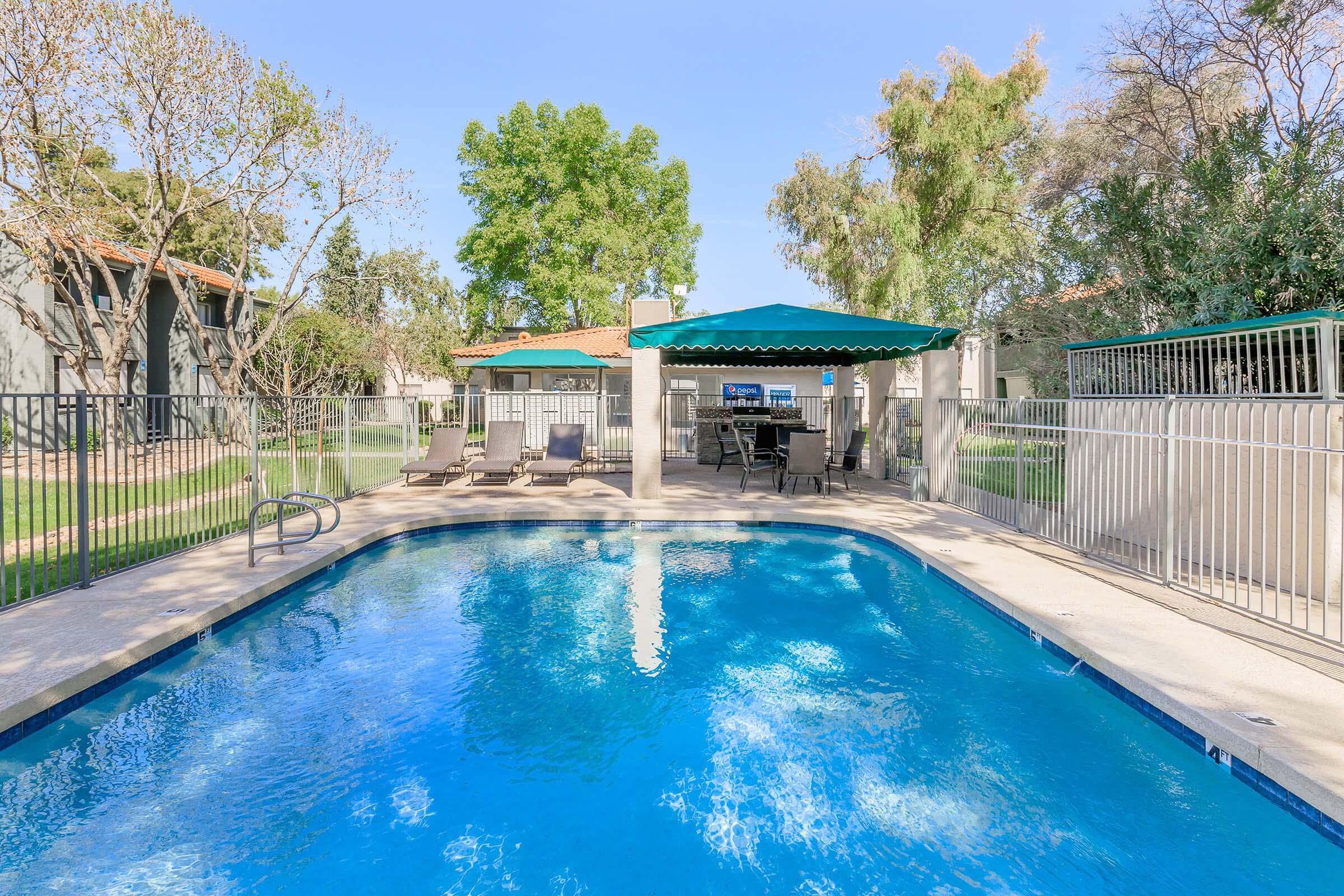 A sunny outdoor pool area featuring a clear blue pool surrounded by lounge chairs, a green shaded gazebo with seating, and well-maintained landscaping. Fenced yard with trees and grass in the background, creating a relaxing atmosphere for leisure and recreation.