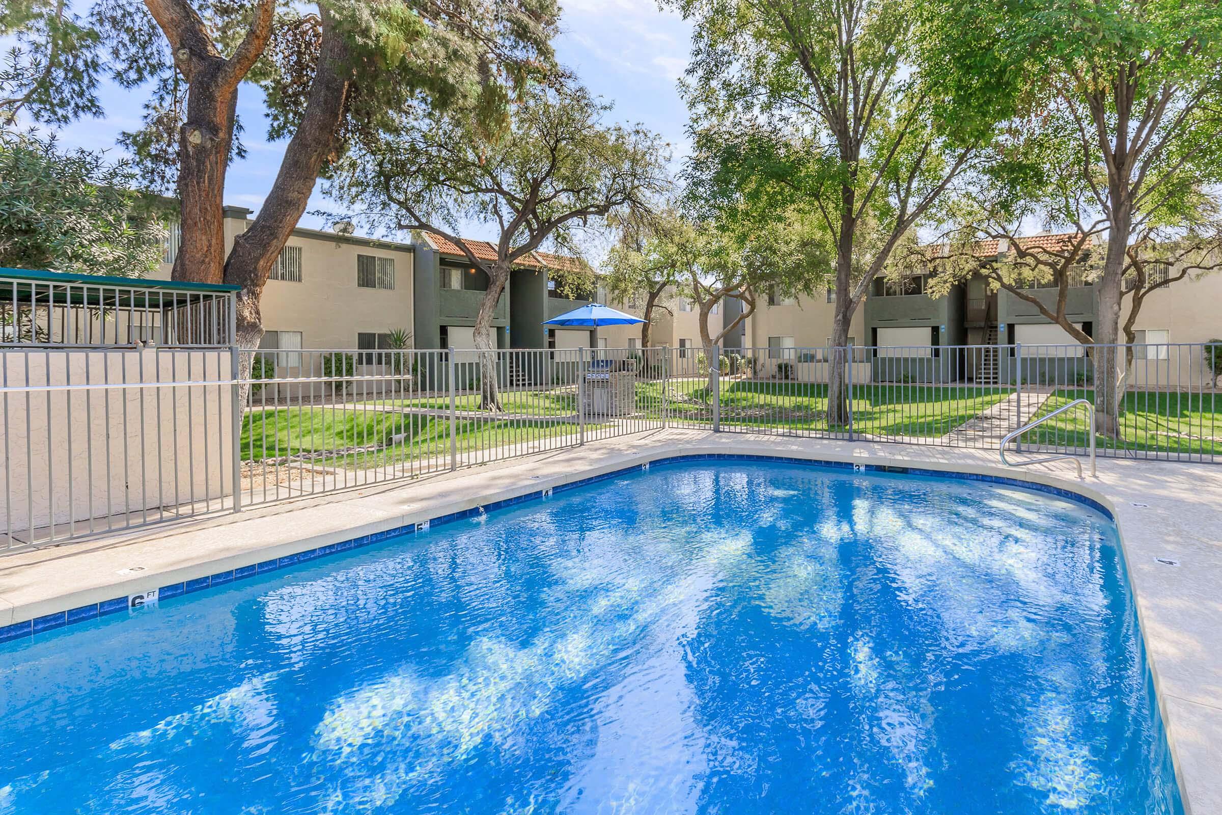 A clear blue swimming pool surrounded by a fence, with green lawns and trees in the background. Apartment buildings are visible behind the pool, providing a residential atmosphere. The scene is bright and sunny, conveying a tranquil outdoor space suitable for relaxation.