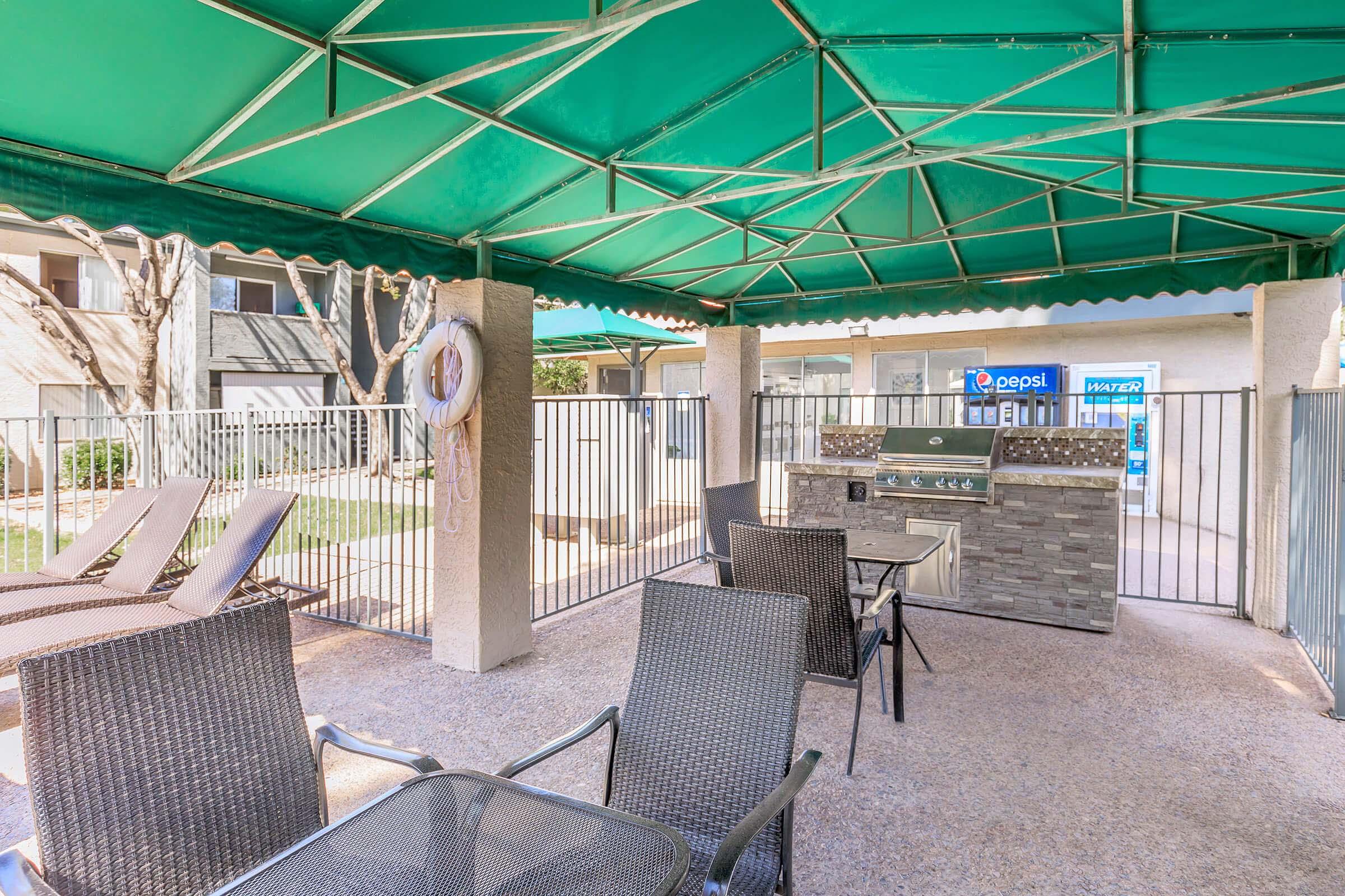 A shaded outdoor area featuring a green canopy, lounge chairs, and a stone barbecue grill. In the background, a fence encloses a pool area, with a vending machine visible. The setting is perfect for relaxation and social gatherings.