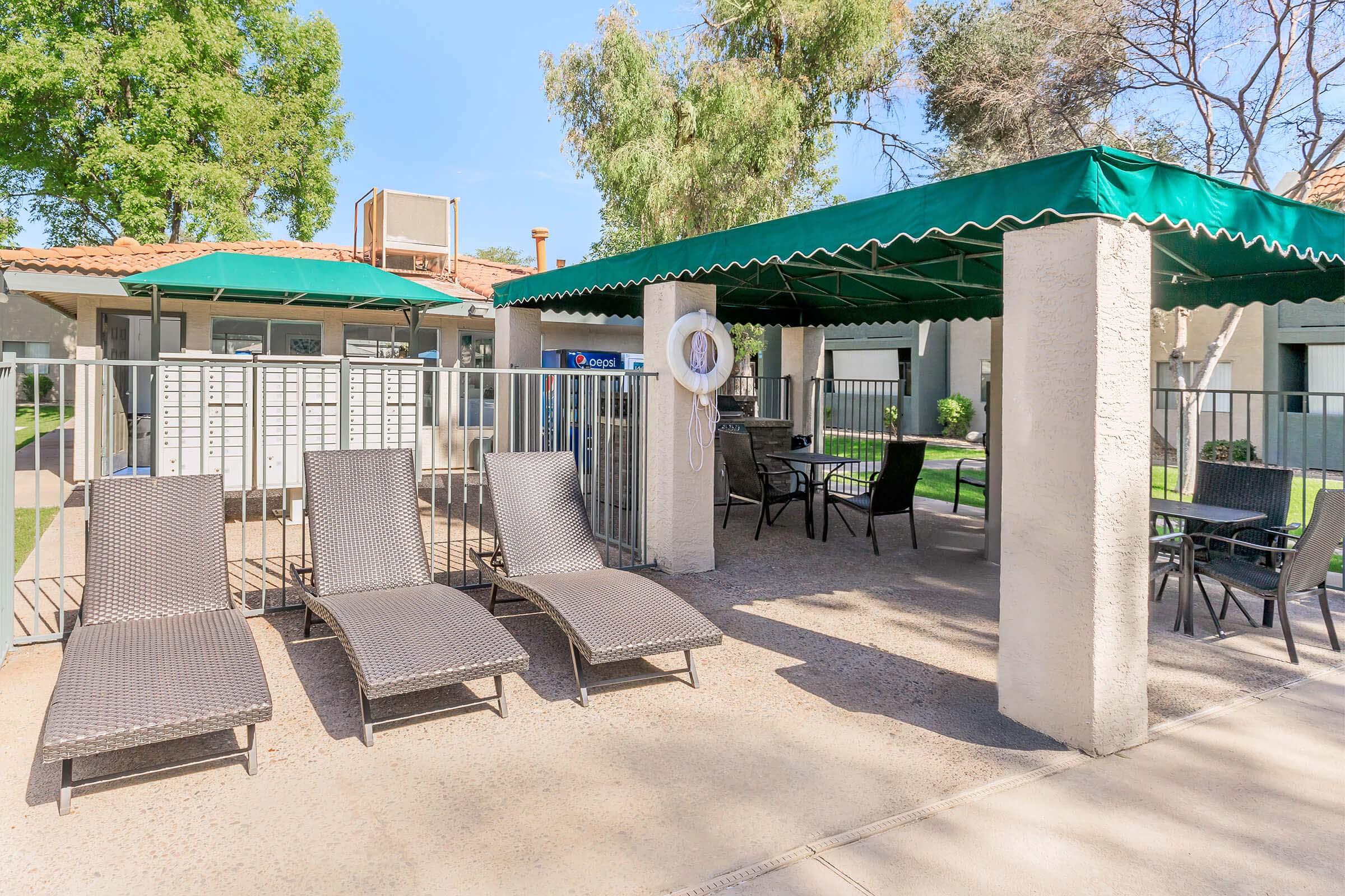 A shaded outdoor area featuring four lounge chairs, a table, and a light fixture, surrounded by a fence. Mailboxes are visible in the background, along with green trees and landscaped grounds, creating a relaxed communal space near an apartment complex.