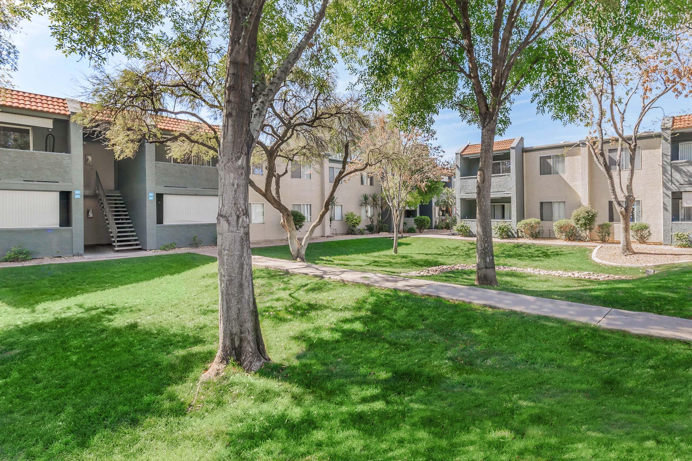 A view of a landscaped outdoor area with green grass and trees, featuring two apartment buildings on either side. The building on the left has a staircase leading to its entrance, and several flowering trees are visible, adding to the pleasant atmosphere of the community.