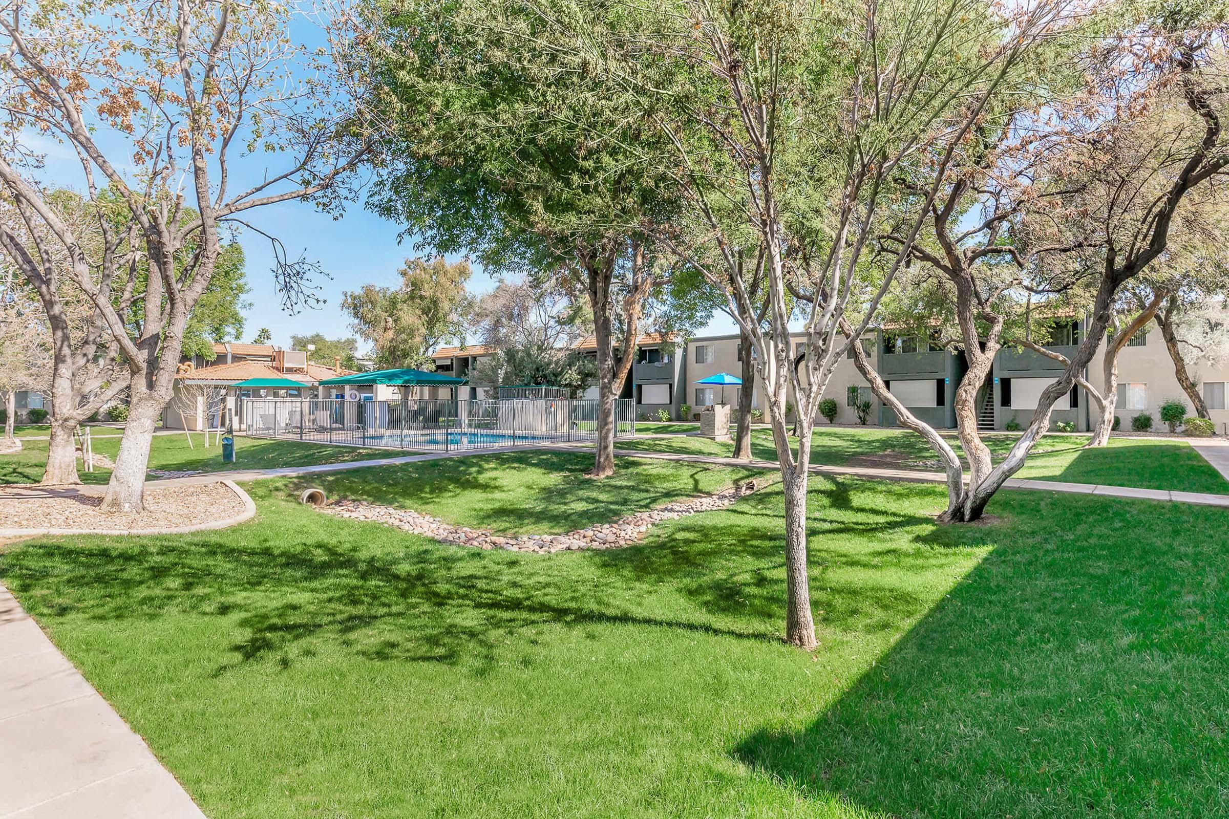 Lush green lawn with trees and pathways, featuring a swimming pool area bordered by lounge chairs and umbrellas. Residential buildings are visible in the background, surrounded by well-maintained landscaping. Bright sunny day with clear blue skies.