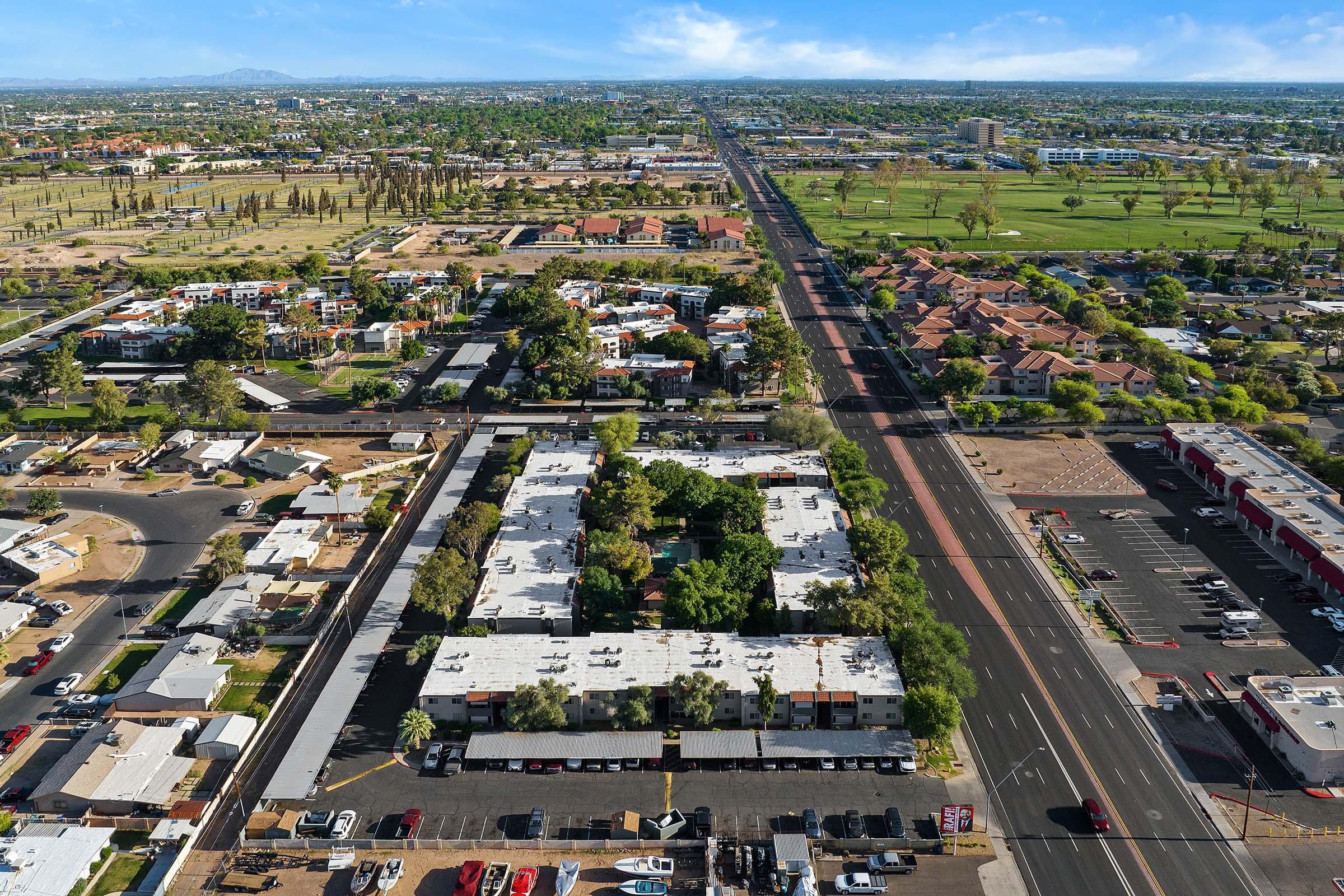 Aerial view of a suburban area featuring residential buildings, a main road lined with trees, and a mix of small commercial spaces and parking lots. The landscape includes green spaces and scattered homes, showcasing a typical urban environment. The scene is bathed in daylight with a clear sky, suggesting a warm climate.