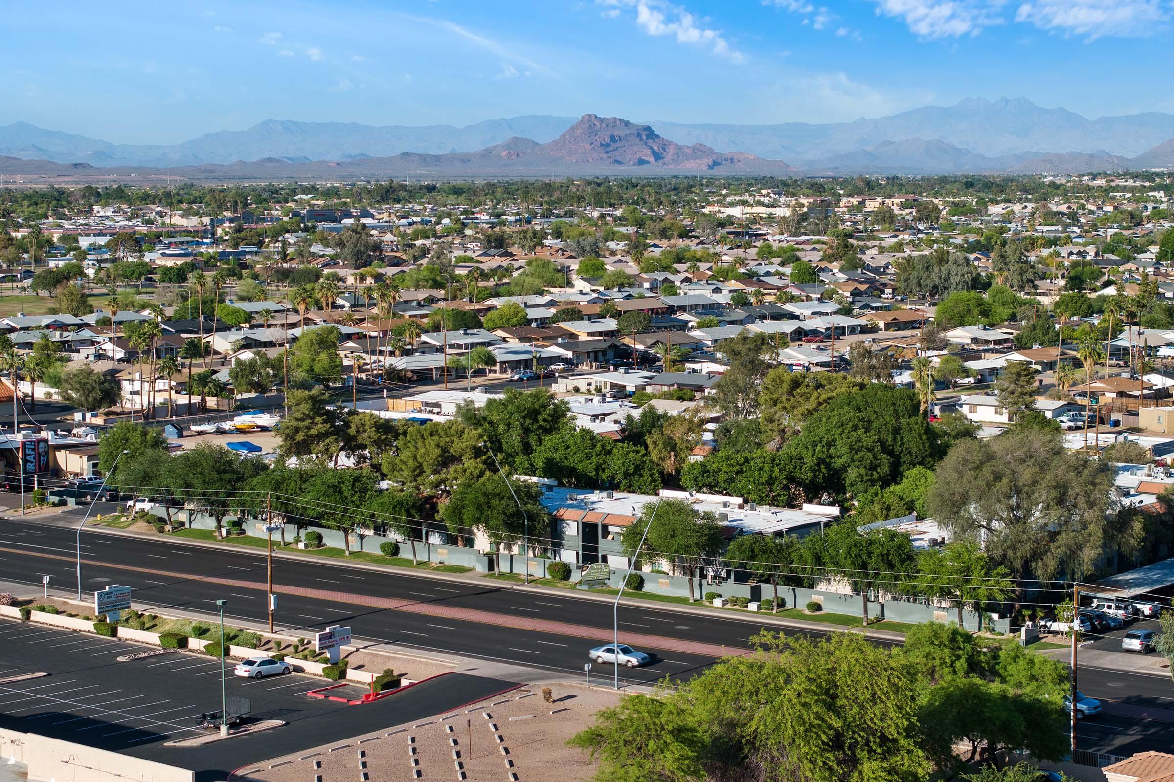 Aerial view of a suburban area featuring densely packed houses, green trees, and a wide road. In the background, mountains rise against a clear blue sky, creating a scenic landscape. The scene showcases a mix of residential buildings and commercial properties, highlighting urban development surrounded by nature.