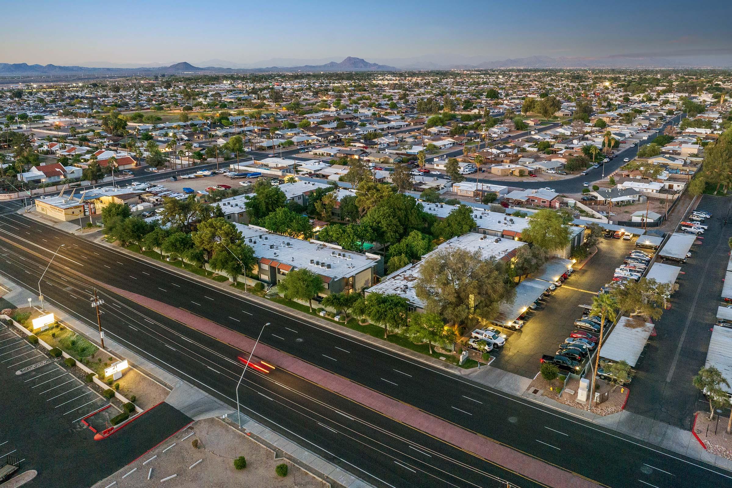 Aerial view of a sprawling suburban area with rows of houses and small commercial buildings. A main road runs along the bottom, lined with parked cars and illuminated parking lots. In the background, mountains are visible under a clear sky, suggesting a tranquil evening atmosphere.