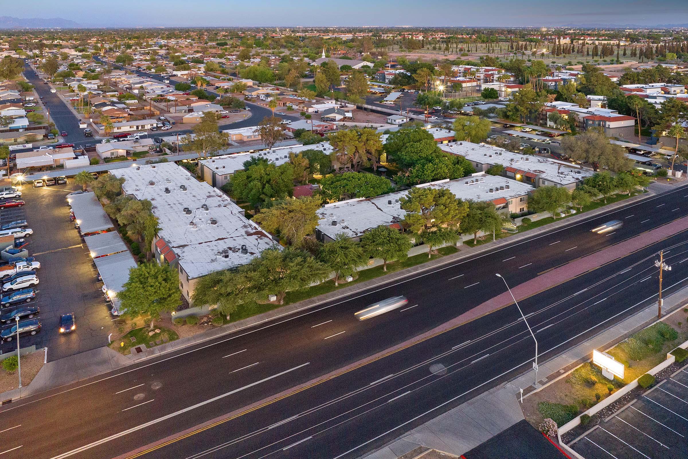 Aerial view of a suburban area featuring a large complex surrounded by greenery, with multiple parking lots and residential buildings. The scene is set during twilight, showing a busy road in the foreground and distant mountains in the background.