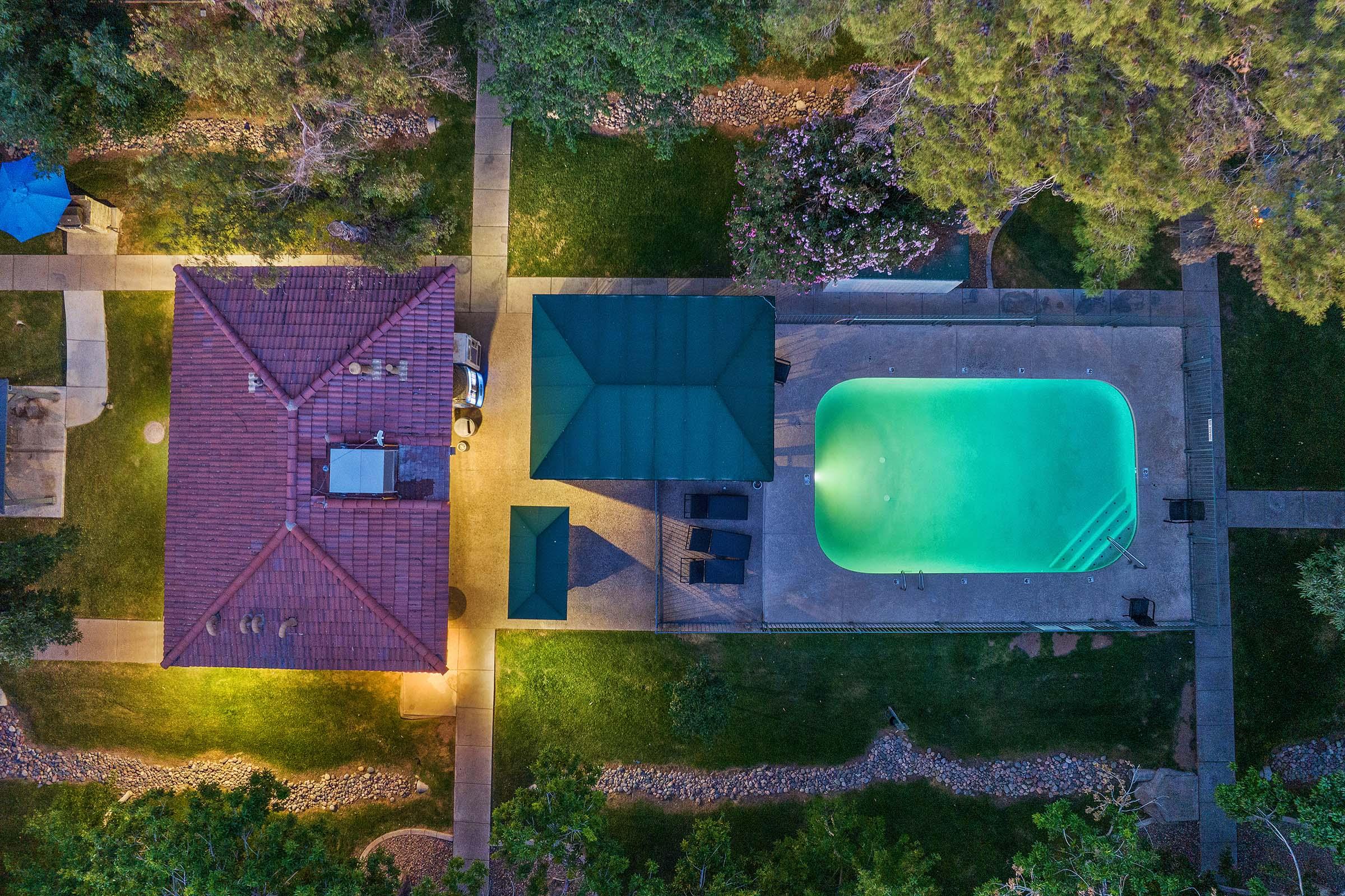 Aerial view of a residential property featuring a house with a red tiled roof, a swimming pool with green-lit water, and shaded seating areas. Surrounding greenery and pathways enhance the outdoor space. The image captures the tranquil atmosphere of a backyard oasis.