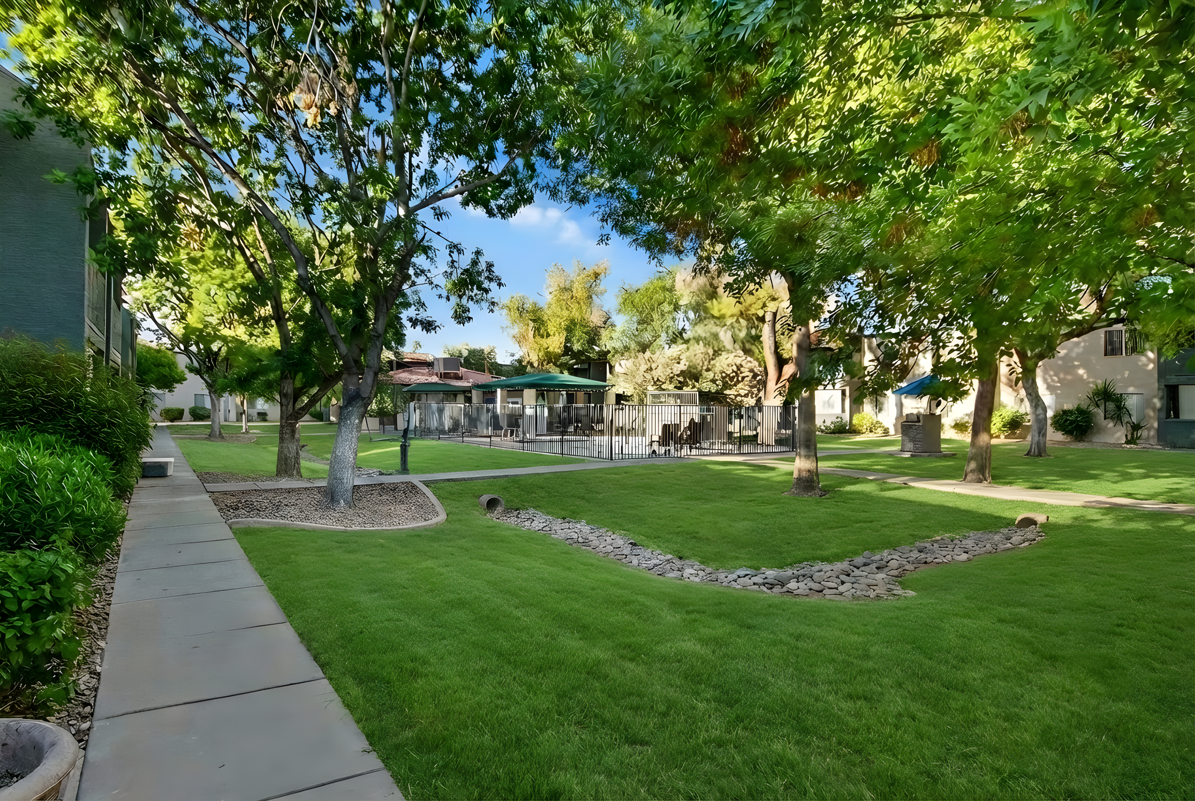 A landscaped area featuring lush green grass, trees, and a winding stone pathway. In the foreground, a path leads through the greenery, while in the background, a fenced pool area is partially visible, surrounded by residential buildings and additional trees under a clear blue sky.