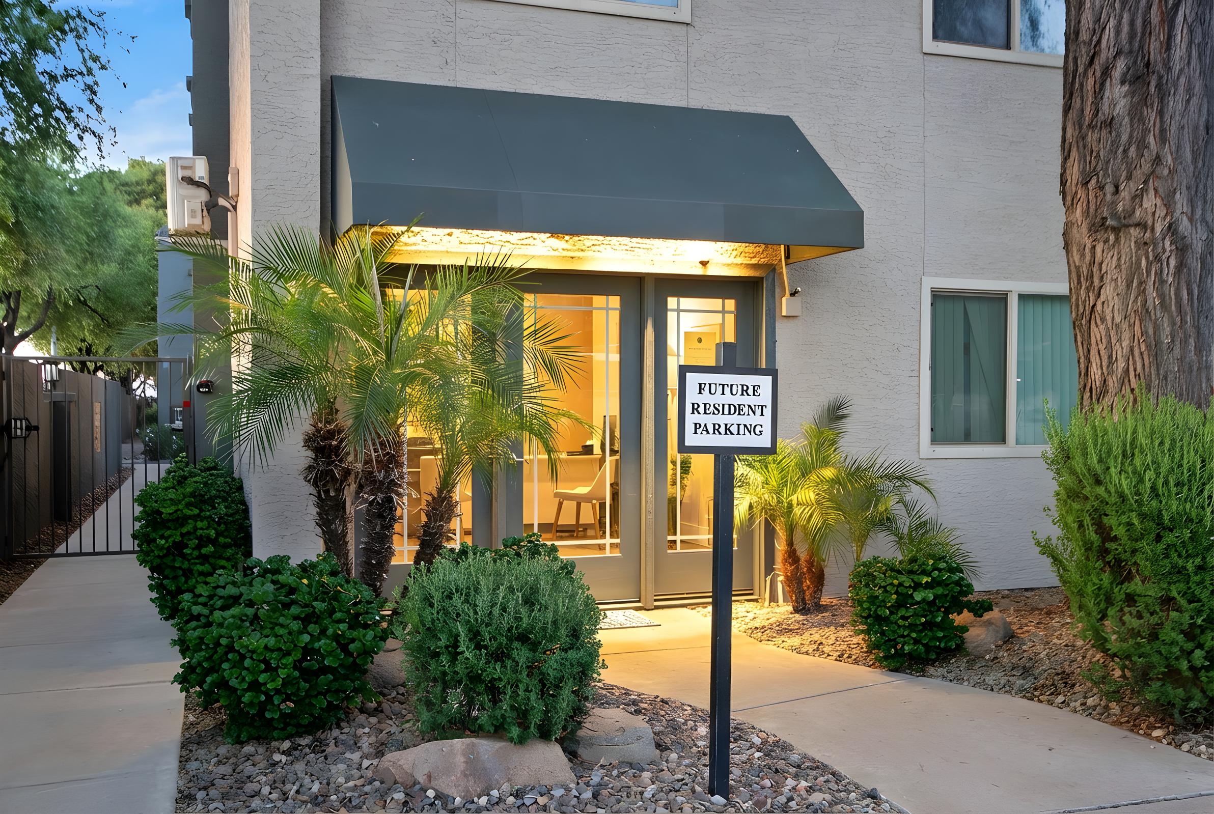 A modern apartment entrance featuring a covered doorway illuminated by warm light. There are potted palm plants on either side of the door, with a sign labeled "FUTURE RESIDENT PARKING" mounted on a post in front. The pathway is lined with small rocks and shrubbery, enhancing the inviting atmosphere.