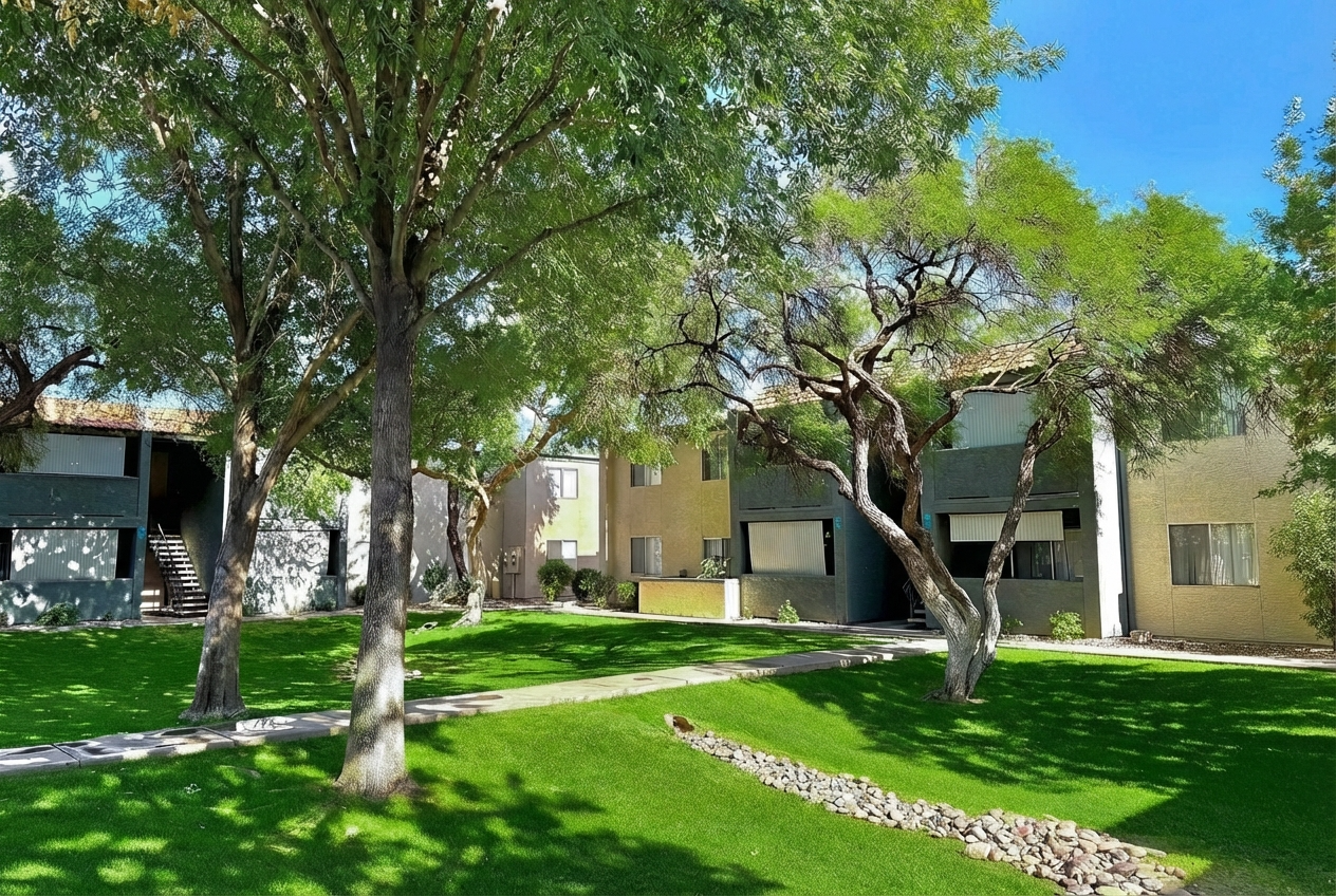 A well-maintained apartment complex surrounded by lush green lawns and trees. The buildings have a modern design with several balconies. A stone pathway runs through the grassy area, providing access to the apartments, all under a clear blue sky.