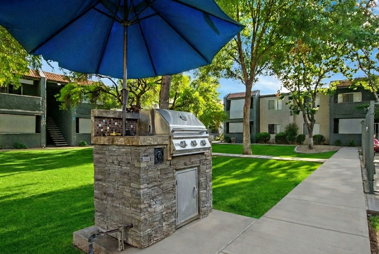 Outdoor barbecue grill with a stone base under a large blue umbrella, situated in a well-maintained green lawn area, surrounded by apartment buildings. The pathway runs alongside the lawn, enhancing the inviting atmosphere of the residential space.