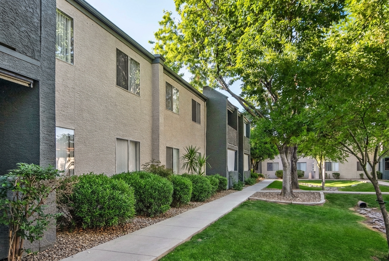 A pathway lined with green bushes leads along a modern apartment building with multiple windows. Trees provide shade on both sides, creating a serene outdoor setting. The scene conveys a well-maintained residential environment.