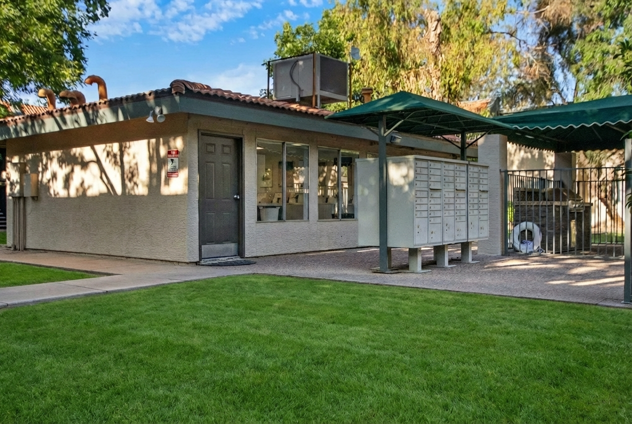 A small, single-story building with a dark door and multiple windows, featuring outdoor mailboxes arranged in a row. The area is surrounded by a green lawn and a fence, with a shaded patio area under a green awning. Trees and blue sky visible in the background.