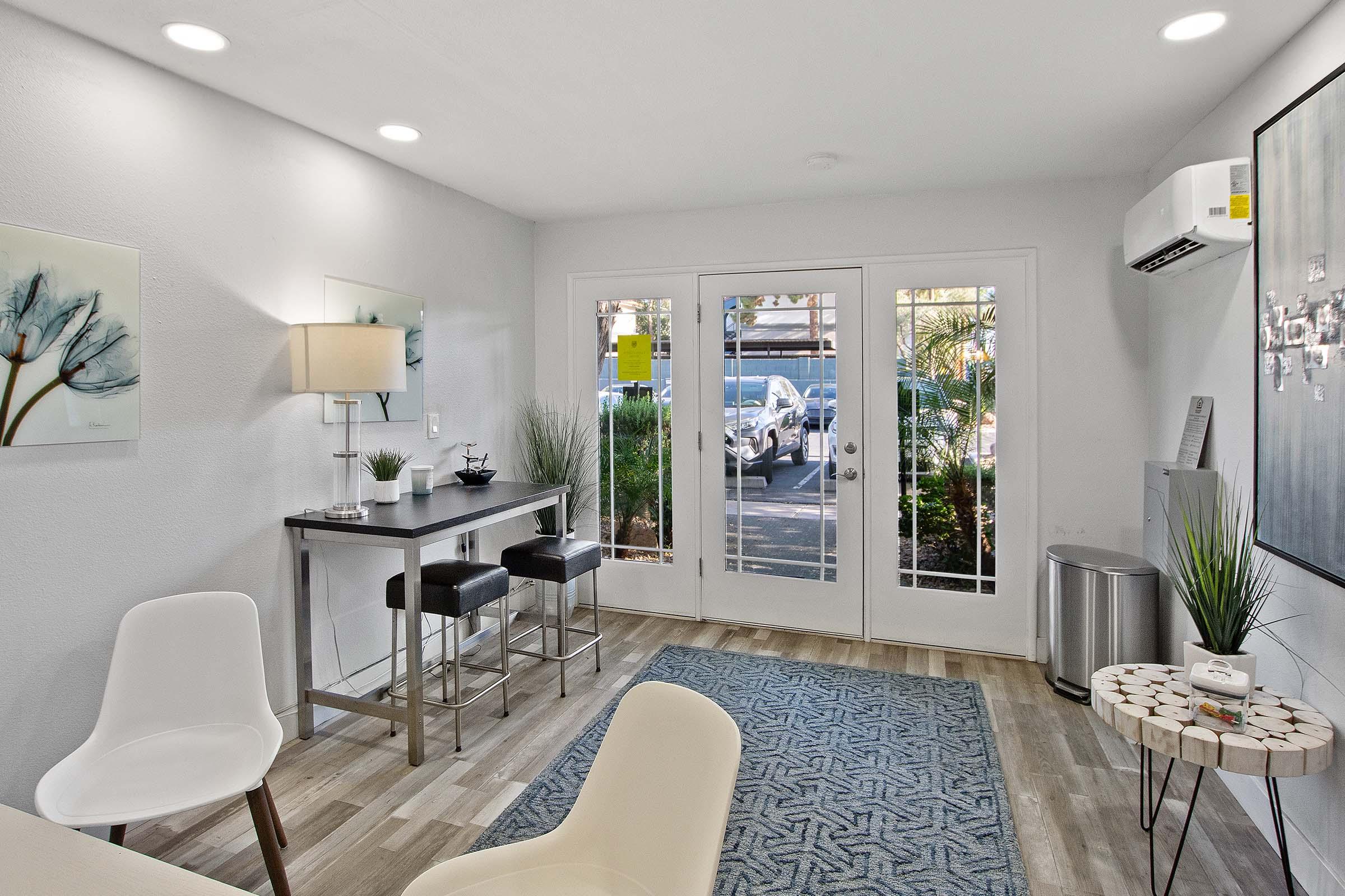 A modern interior view of a bright room featuring a small dining table with two stools, a decorative lamp, and two white chairs. Large doors with glass panels lead outside, revealing greenery. A decorative rug on the floor adds texture to the space, which has neutral wall colors and contemporary decor.