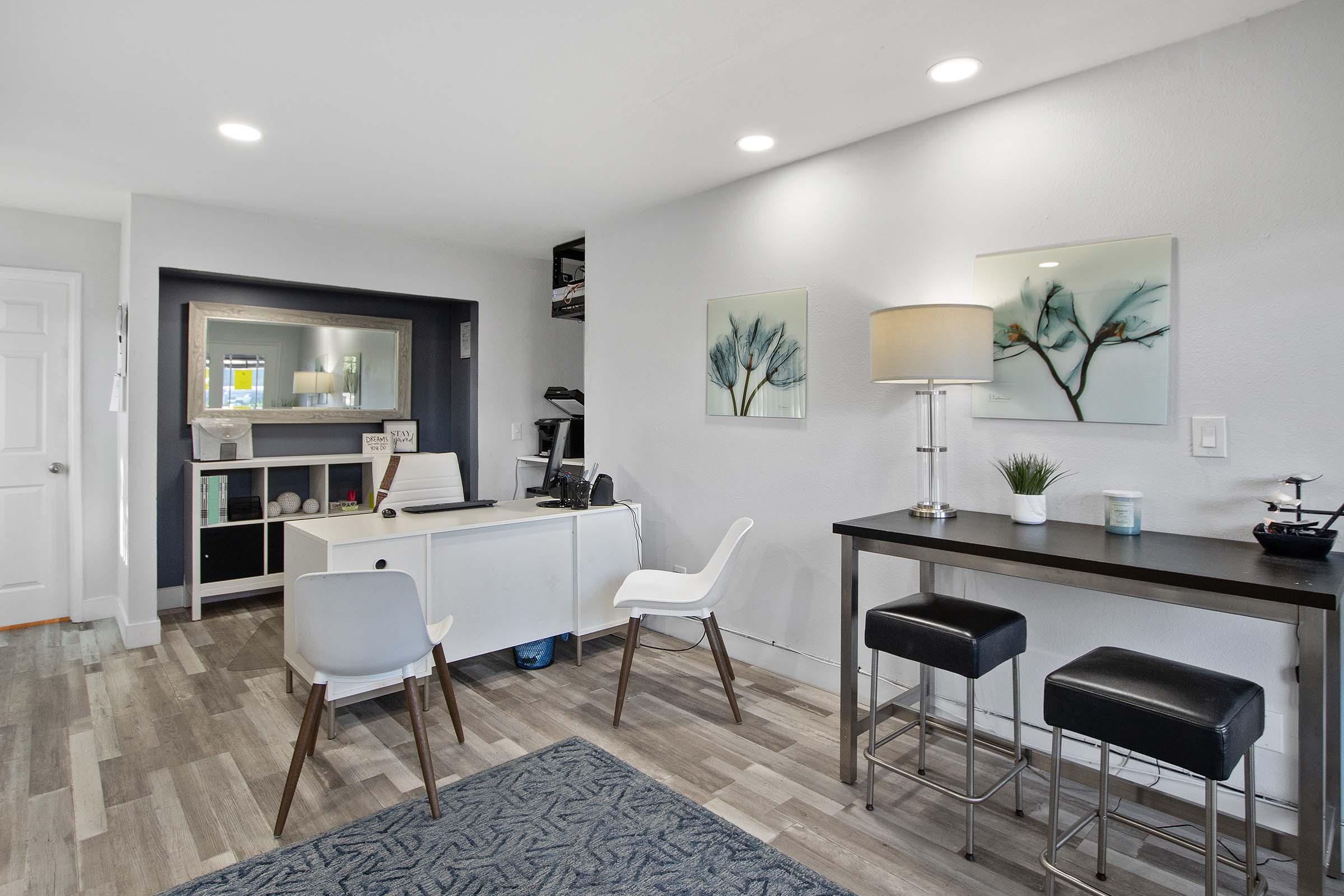 A modern office space featuring a white desk with a computer, two stylish white chairs, and a small black bar table with stools. The walls are adorned with blue-tinted artwork, and there is a lamp on the desk. The flooring is wood-like, and the room is well-lit with recessed lighting.