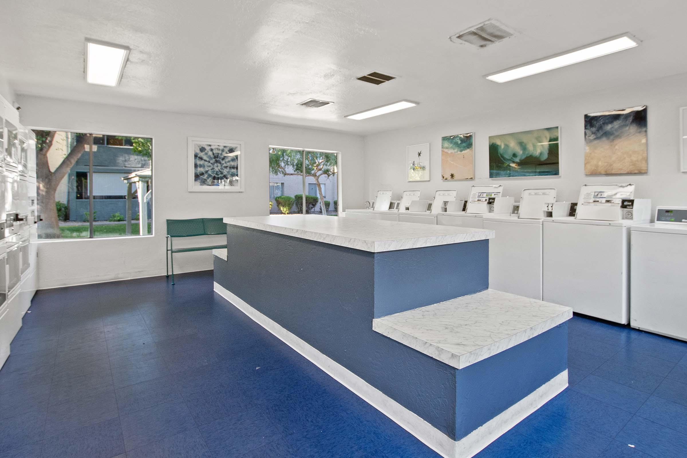 A clean, modern laundromat interior featuring white washing machines and dryers along the wall. A central marble countertop with a small seating area and large windows allowing natural light to illuminate the space. The walls are adorned with abstract paintings, creating a welcoming atmosphere.