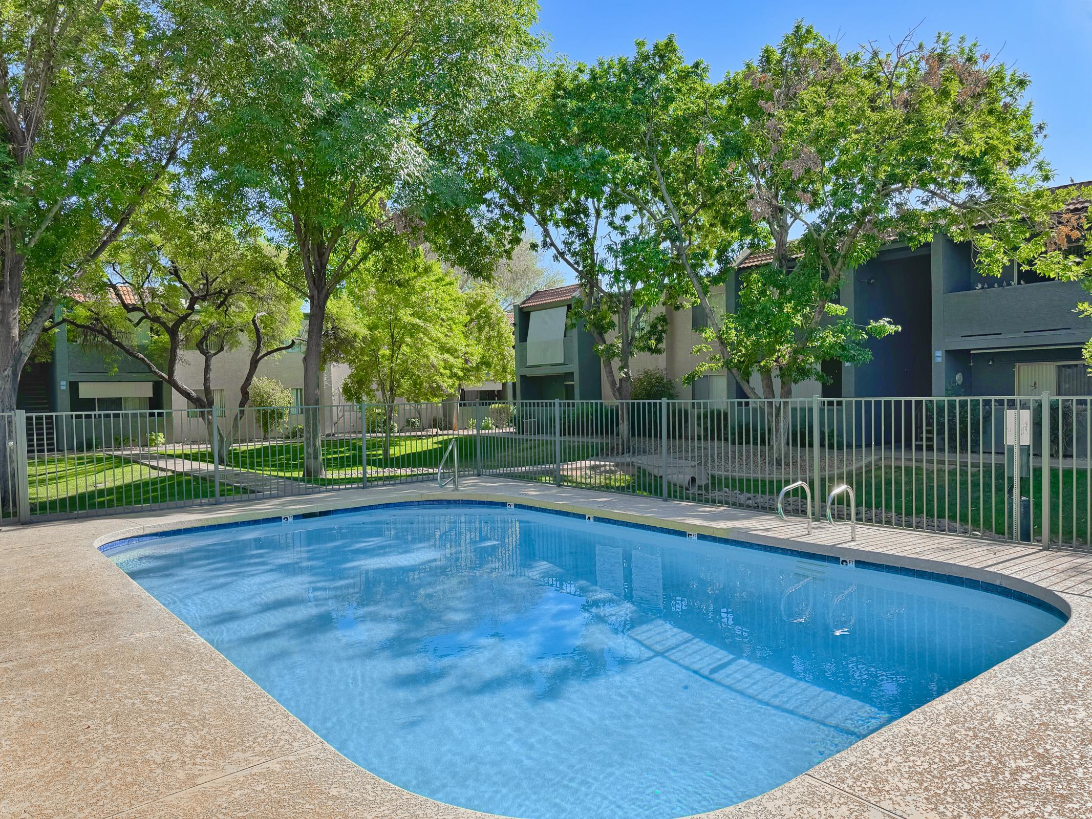 A clear, well-maintained swimming pool surrounded by green trees and grassy areas, with modern apartment buildings in the background. The pool has a fence for safety and is bathed in bright, sunny weather.