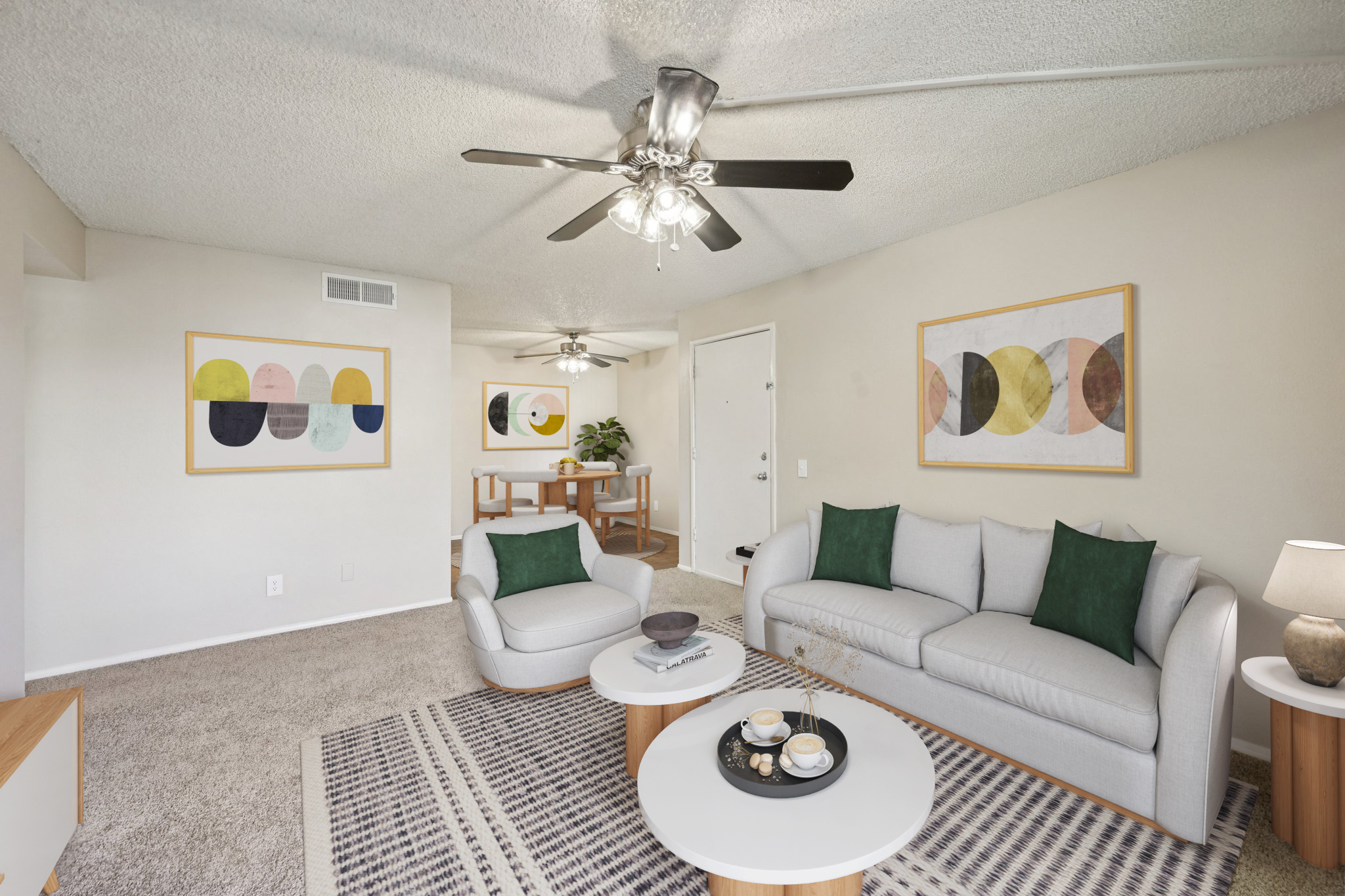 A cozy living room featuring a light gray sofa with green accent pillows, a coffee table with decorative items, and a patterned rug. There are modern art pieces on the walls, a dining area in the background, and ceiling fans providing a comfortable atmosphere.