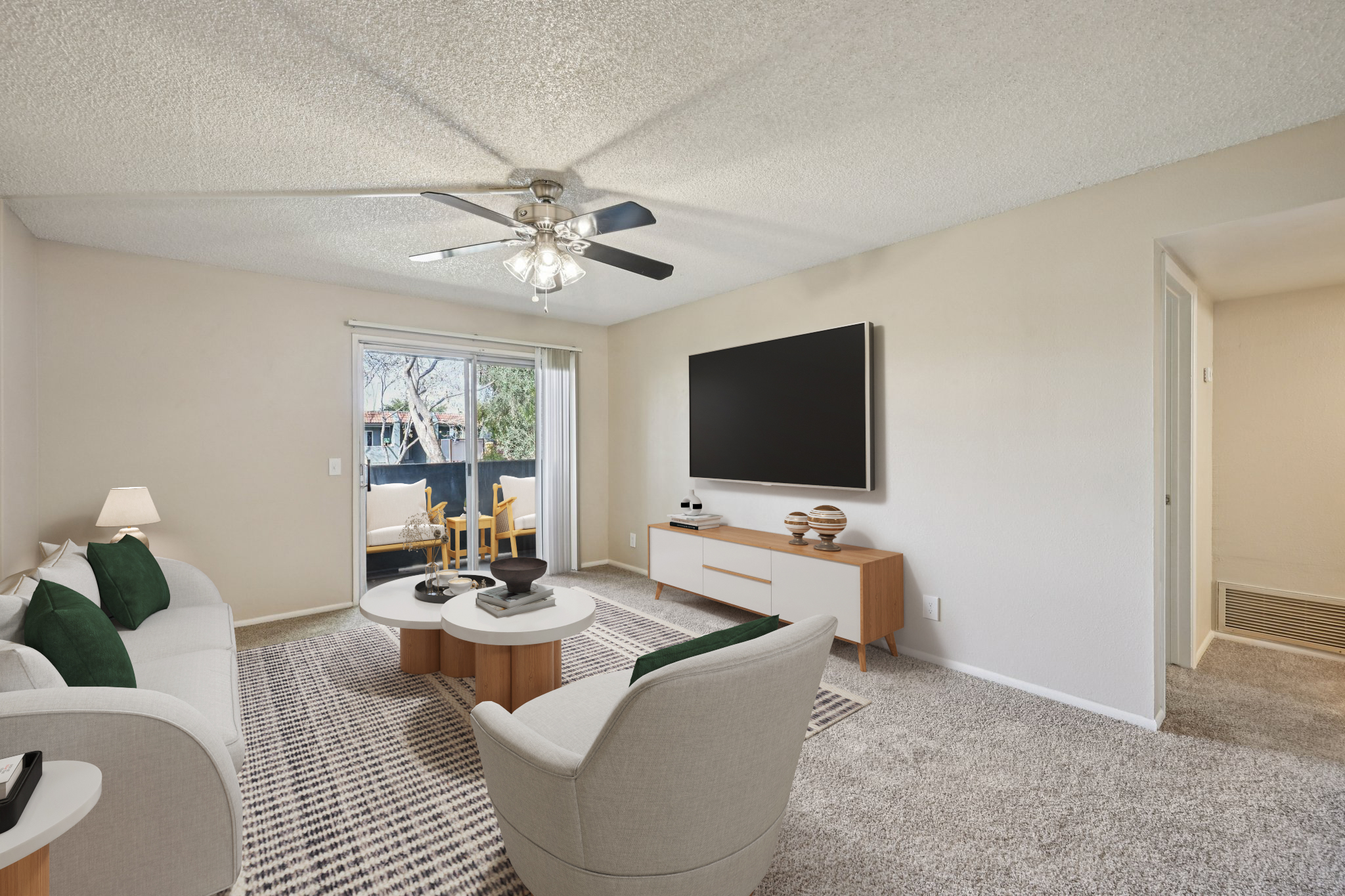 A modern living room featuring a light-colored couch with green cushions, a circular coffee table, and an entertainment unit with a large flat-screen TV. There is a ceiling fan above, and a warm natural light comes through a sliding glass door, leading to a small balcony with outdoor seating.